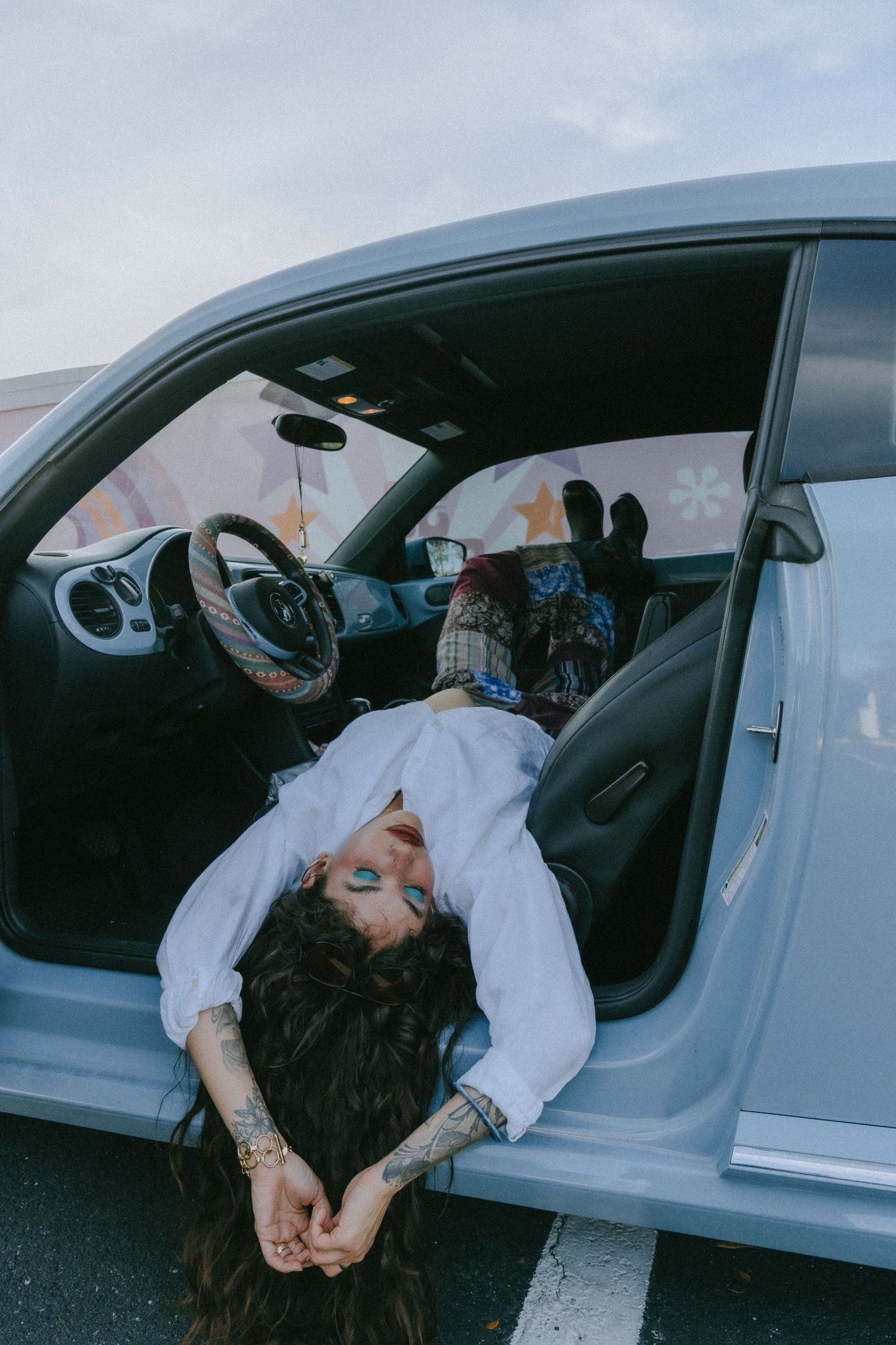 Tattooed woman in white shirt lying across car seat with door open, blue eye makeup, dark hair flowing down.