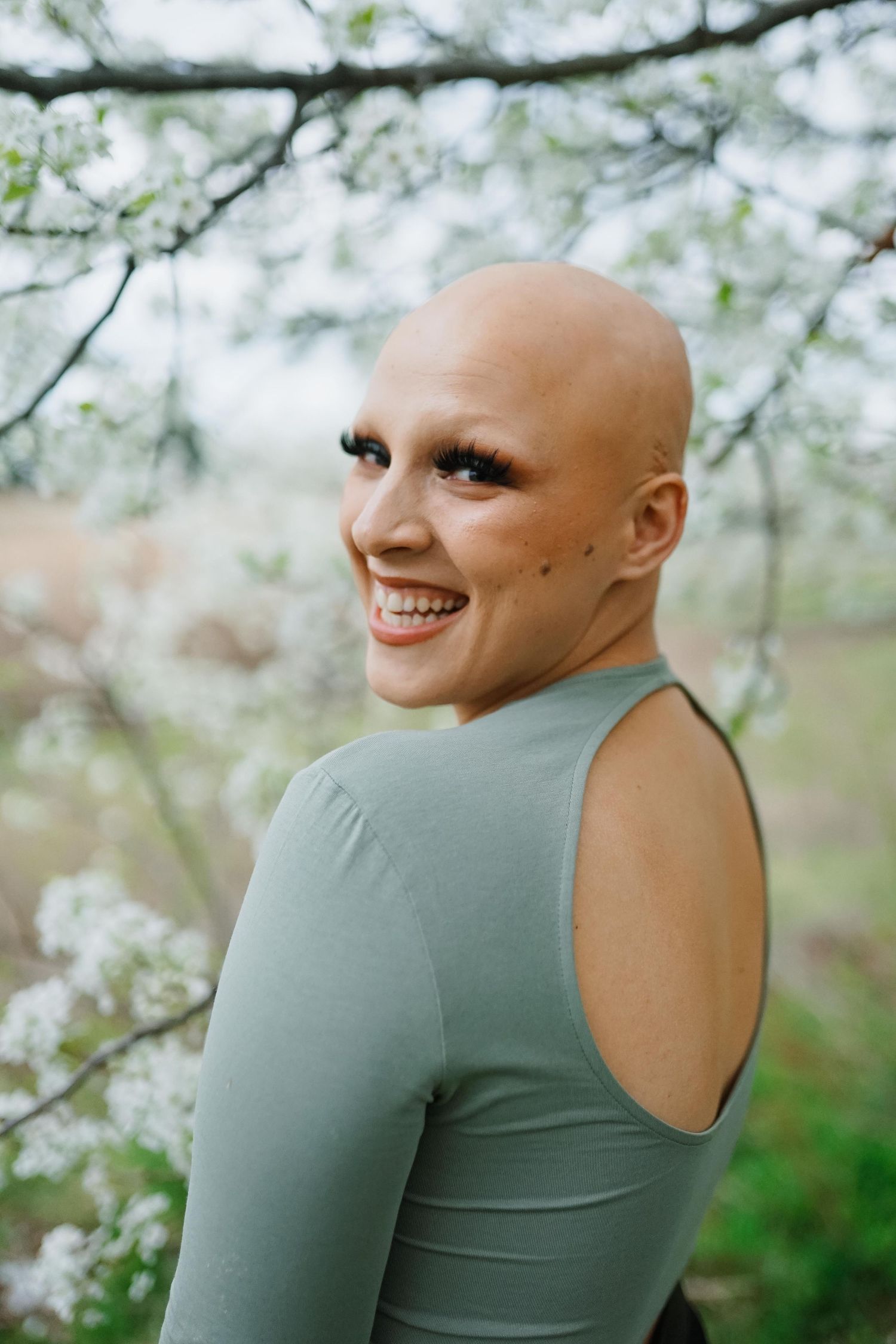 Someone wearing a blue-gray cold shoulder top smiles in front of blooming white spring trees on a soft-focus background.