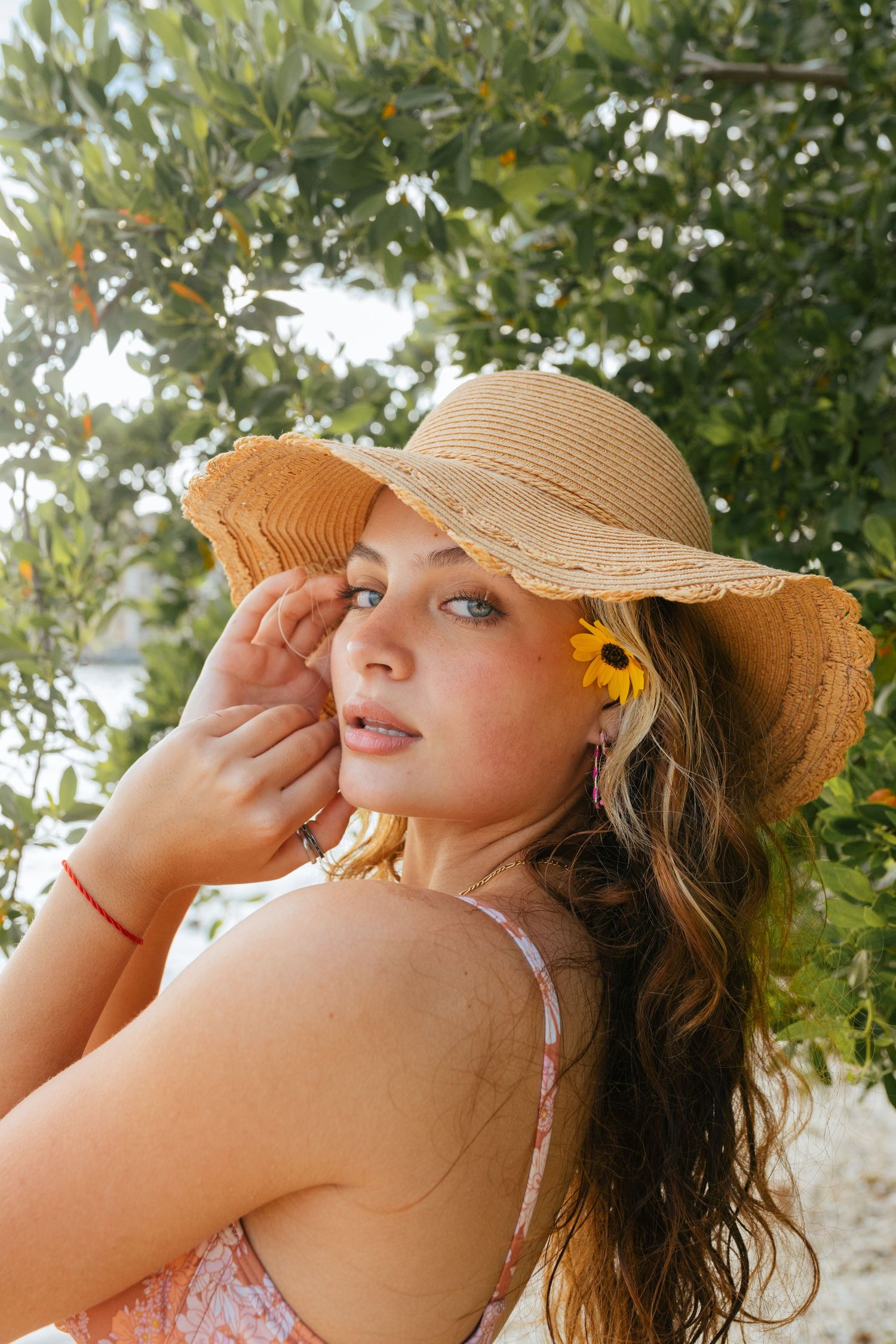 Woman in straw hat with sunflower earring poses thoughtfully against green foliage backdrop.