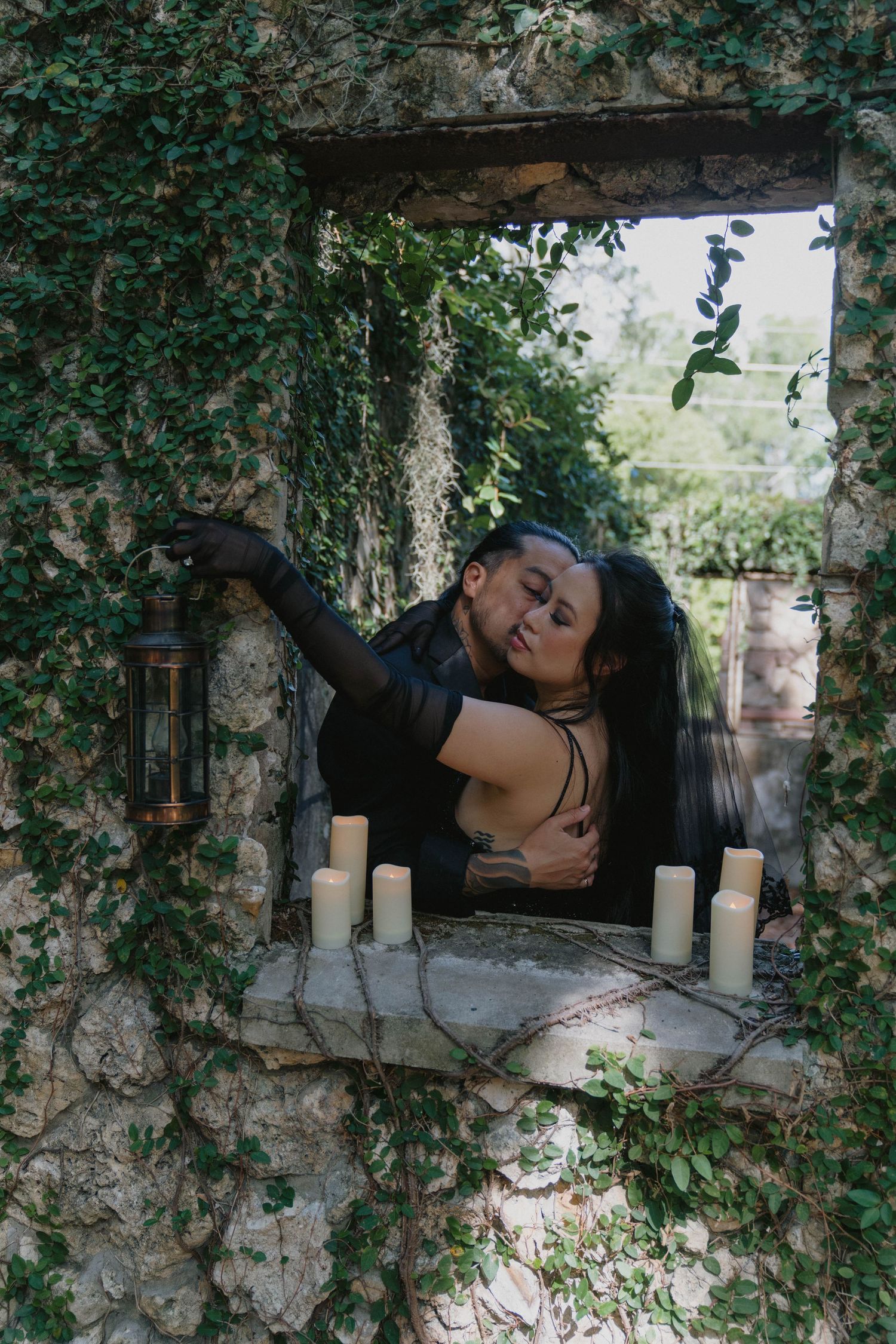 Couple in dark gothic attire embracing in ivy-covered stone window with candles, romantic outdoor fantasy setting.