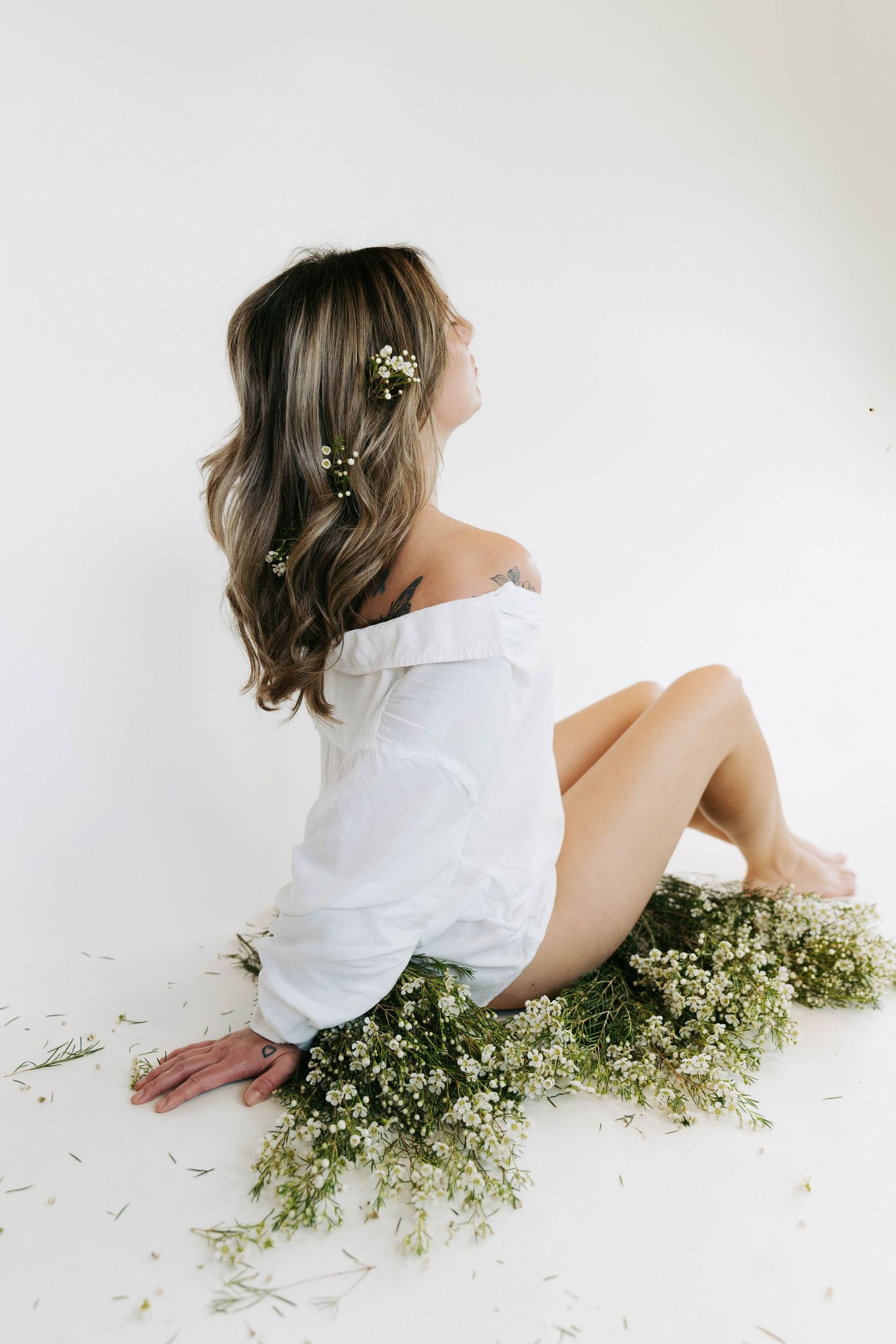 Woman in white off-shoulder top sits on floor surrounded by wildflowers with small blooms in her wavy hair.