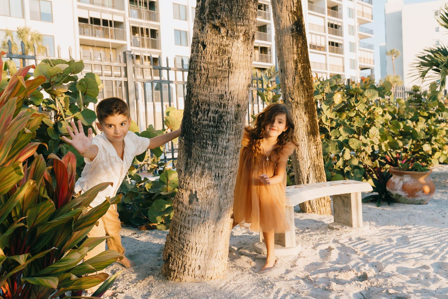 Two children playing hide and seek around a palm tree on a sandy beach with tropical plants and buildings.