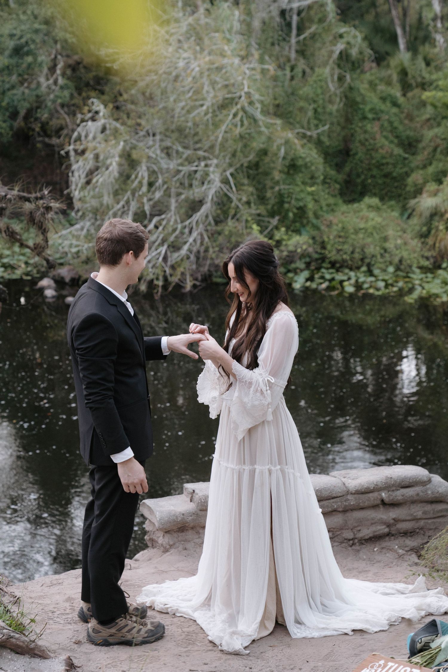 Couple in formal attire shares romantic moment by a serene pond in a natural outdoor setting.
