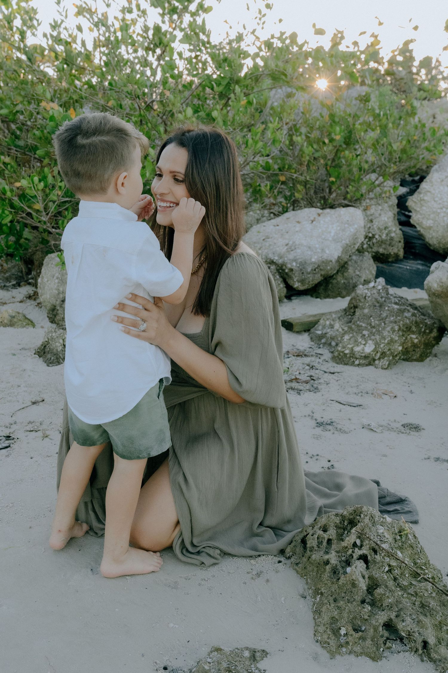A tender moment shared between two people on a sandy beach during sunset with rocks and greenery in the background.