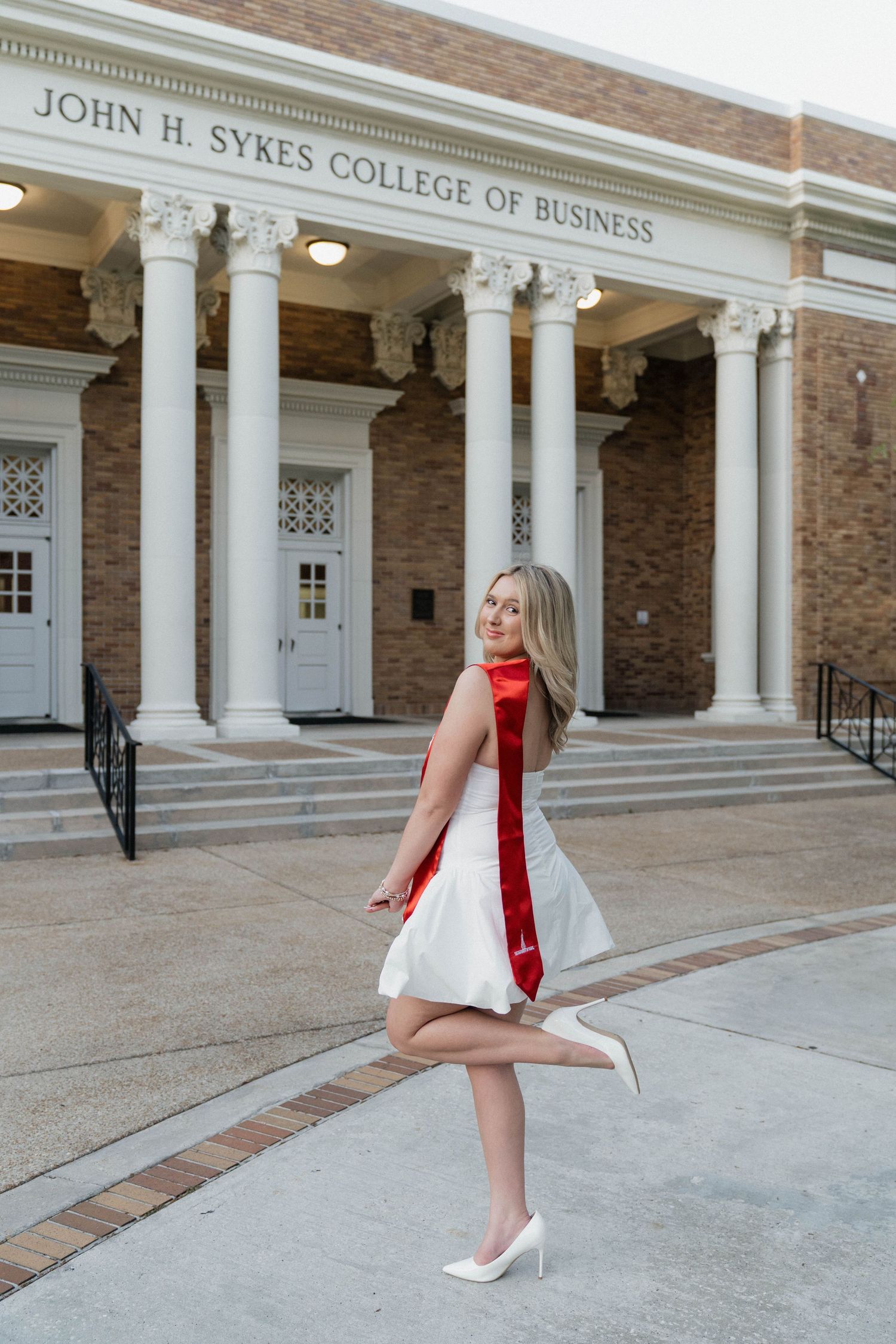 Smiling graduate in white dress and red sash poses playfully outside John H. Sykes College of Business.