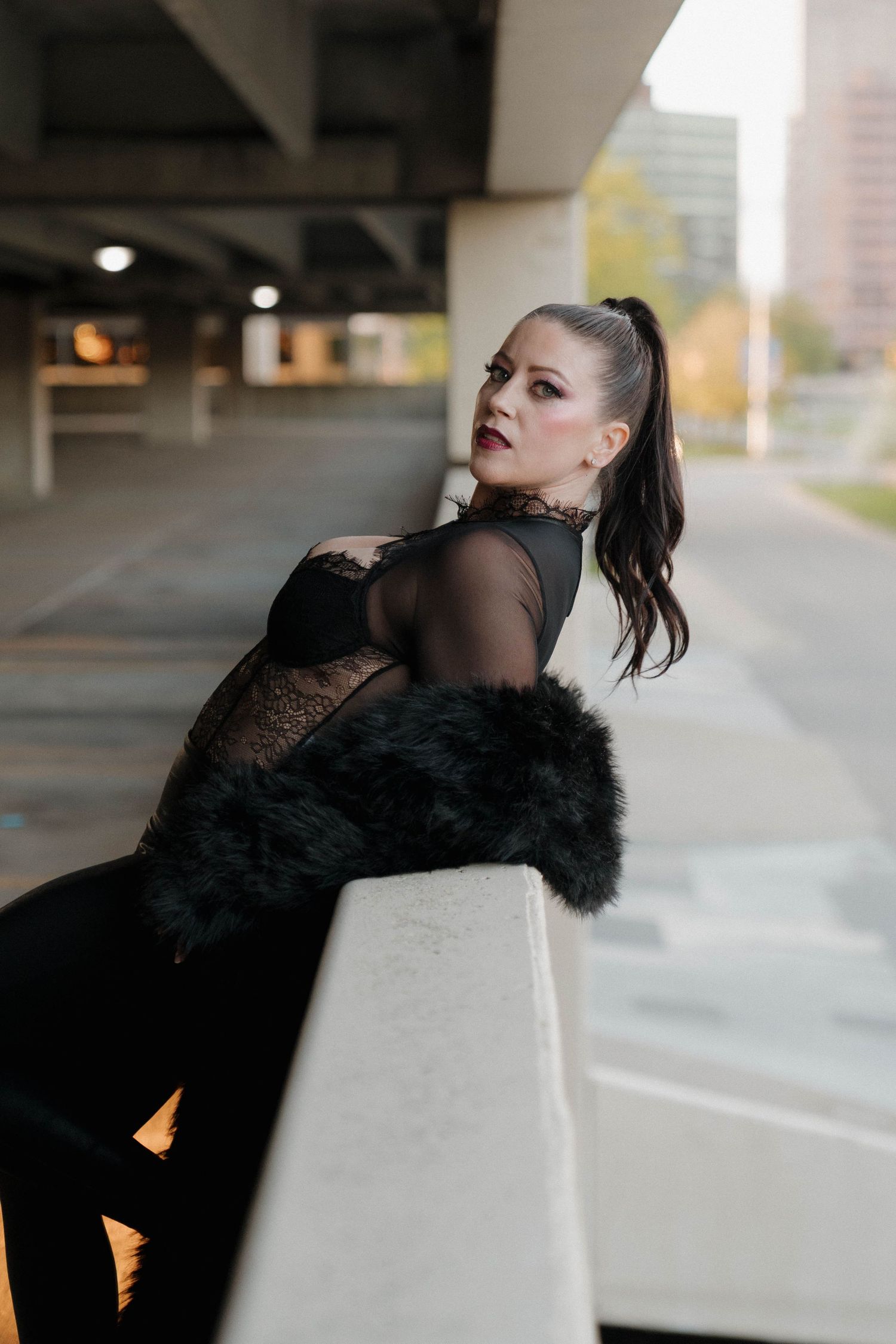 Woman in black lace bodysuit and fur coat poses against concrete barrier in urban parking structure.
