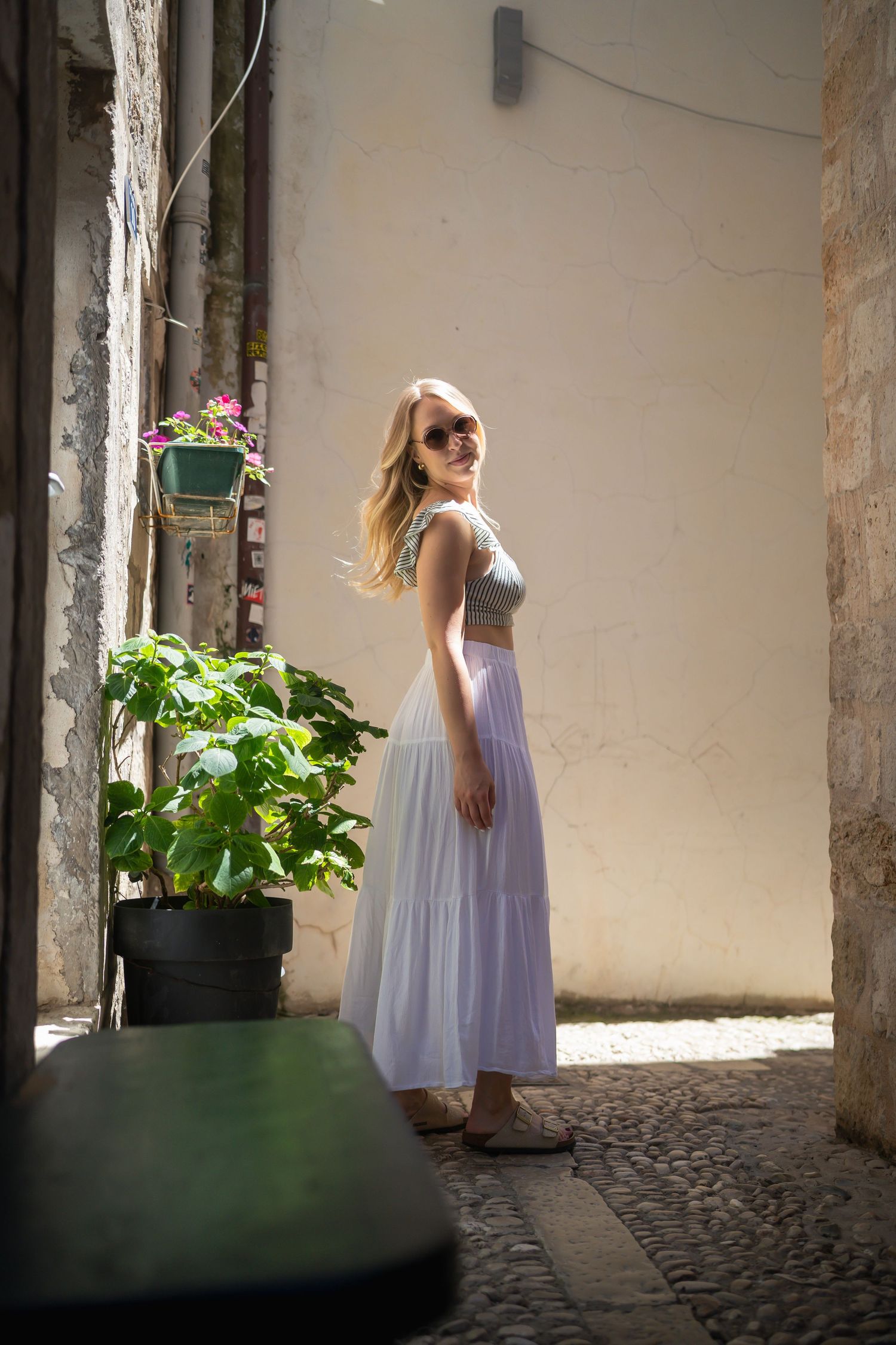 travel photo of beautiful blonde woman standing in a sliver of sunlight looking back to camera in dubrovnik, croatia cobblestone alleyway