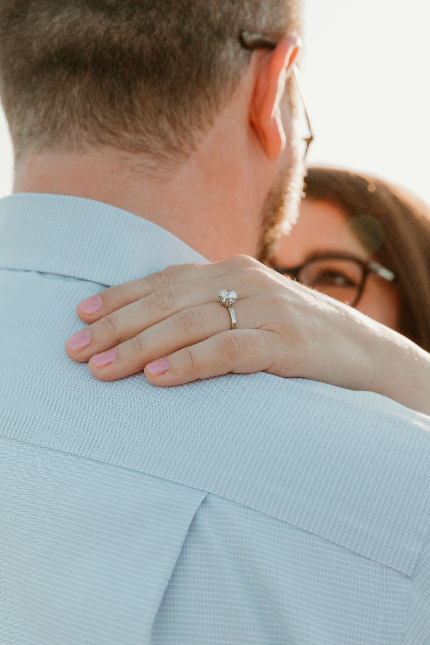 Close-up of hands with engagement ring resting on shoulder in soft lighting.
