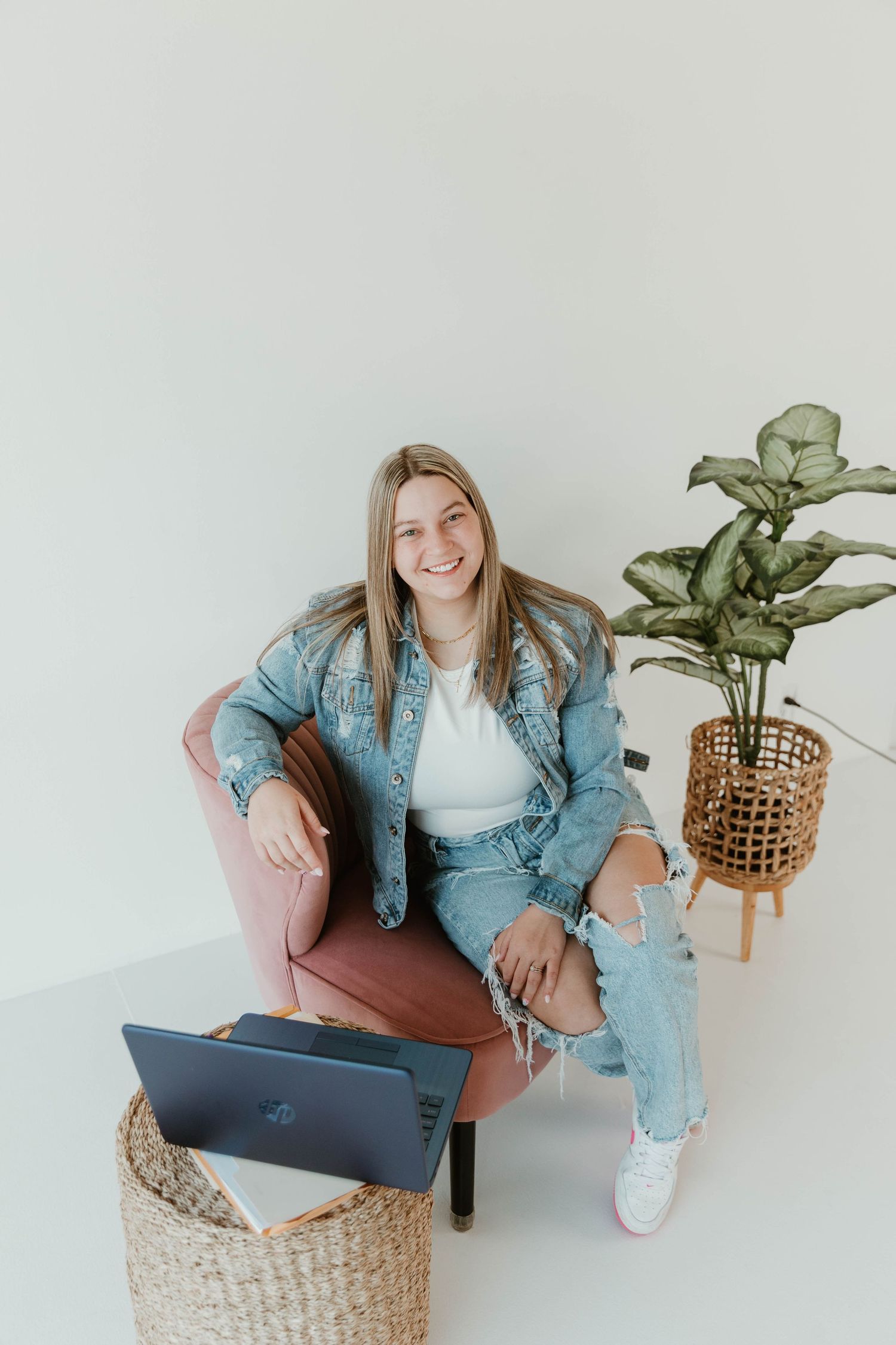 Person seated in brown chair wearing denim outfit with laptop and woven basket nearby.