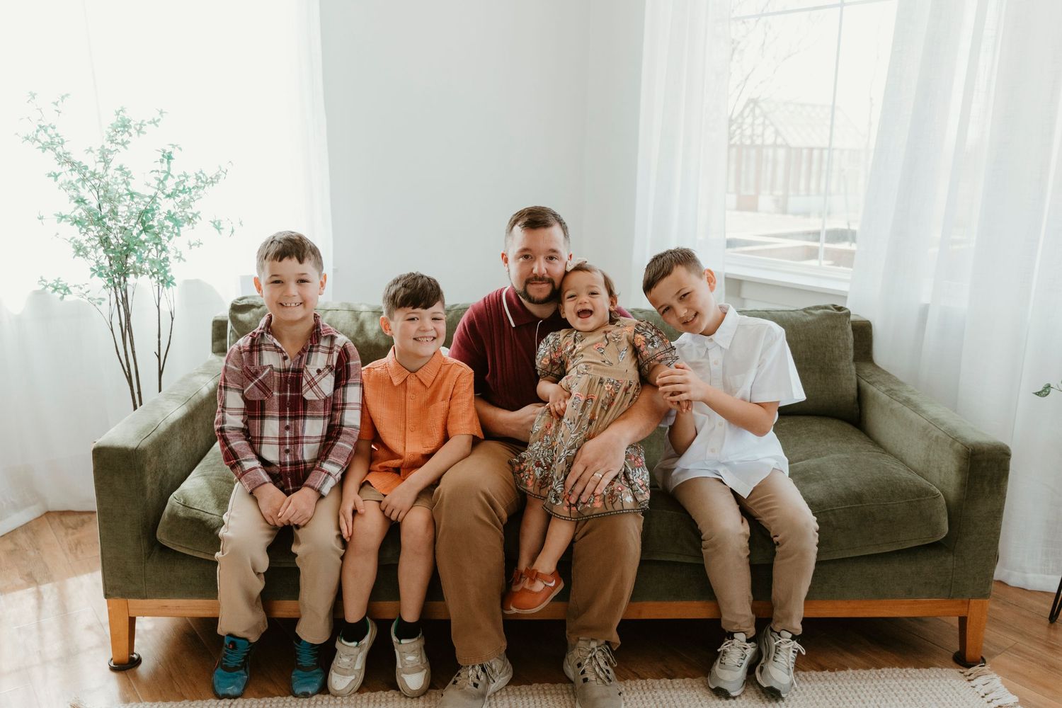 Modern family portrait with five people seated on olive green couch in sunlit room.