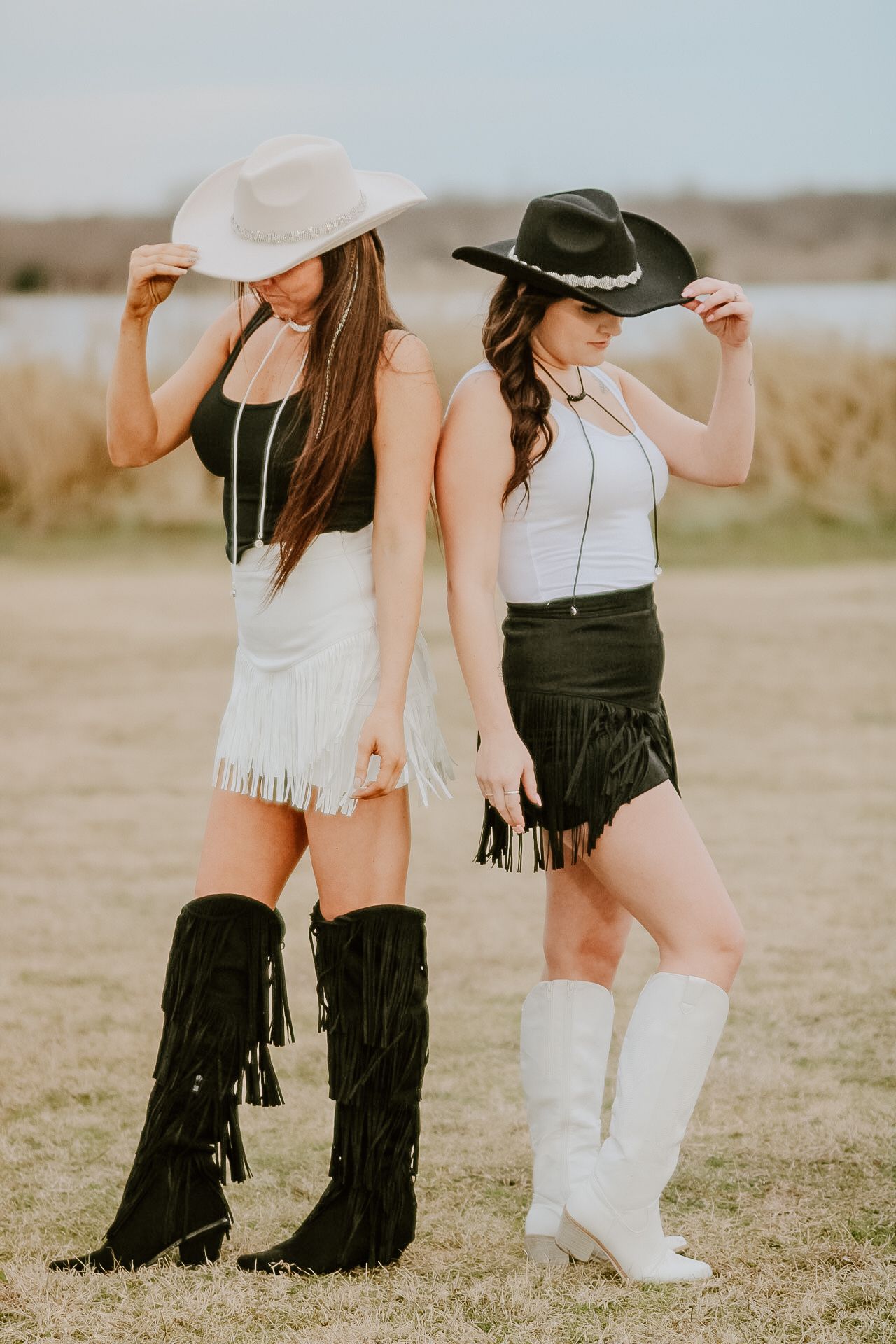Two women in cowgirl outfits with fringe skirts and boots posing in a field at sunset.