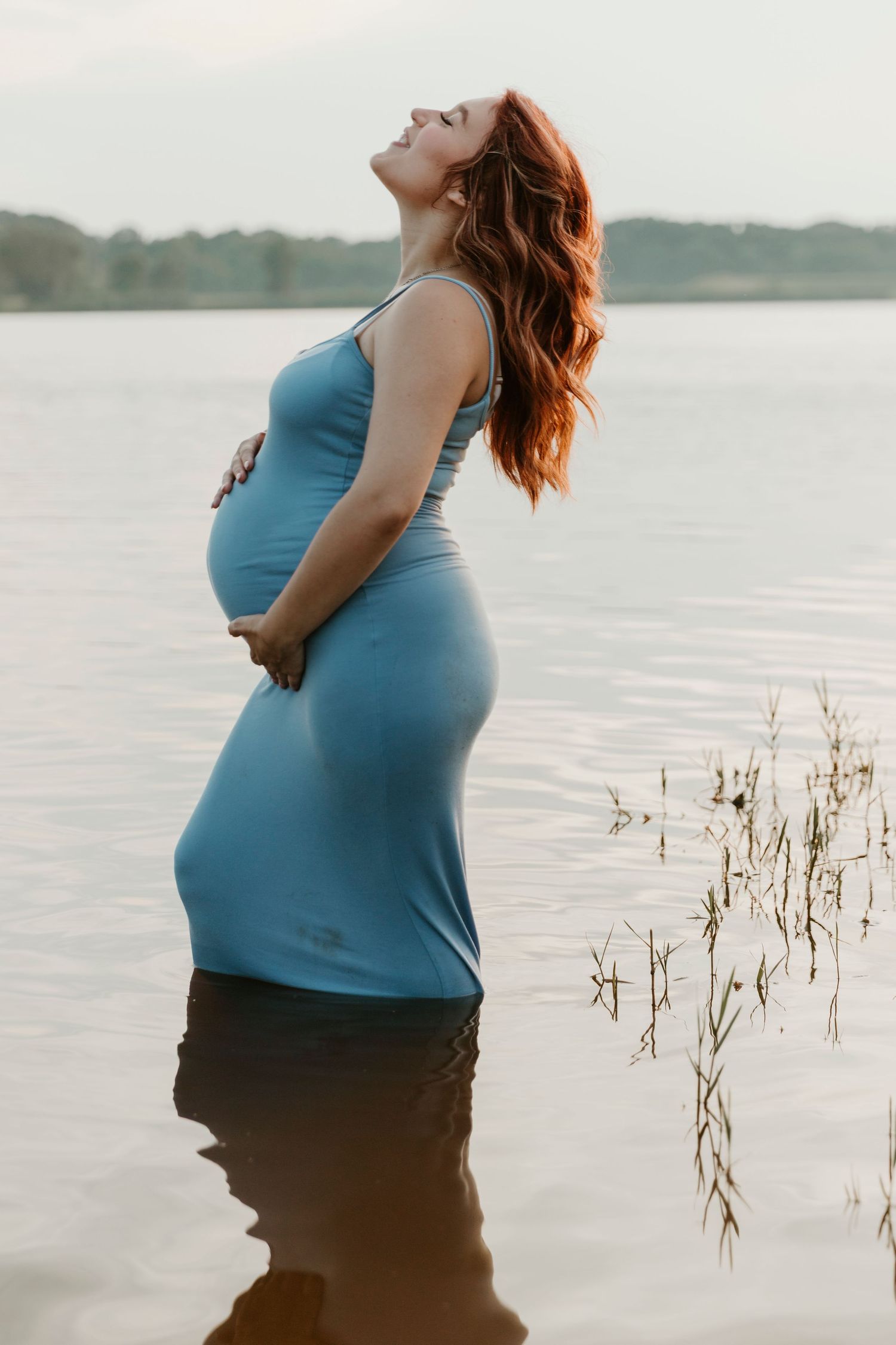 Maternity photo of woman in blue dress standing in water with sunlit lake backdrop.