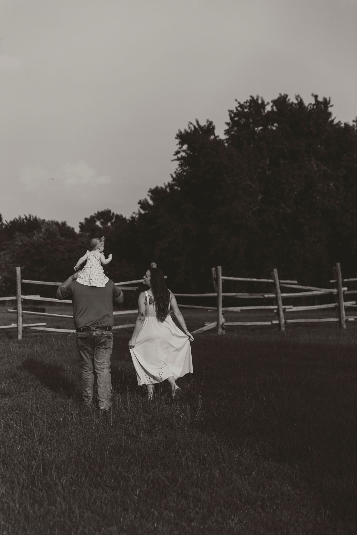 A couple walks through a field at dusk with a wooden fence in the background.