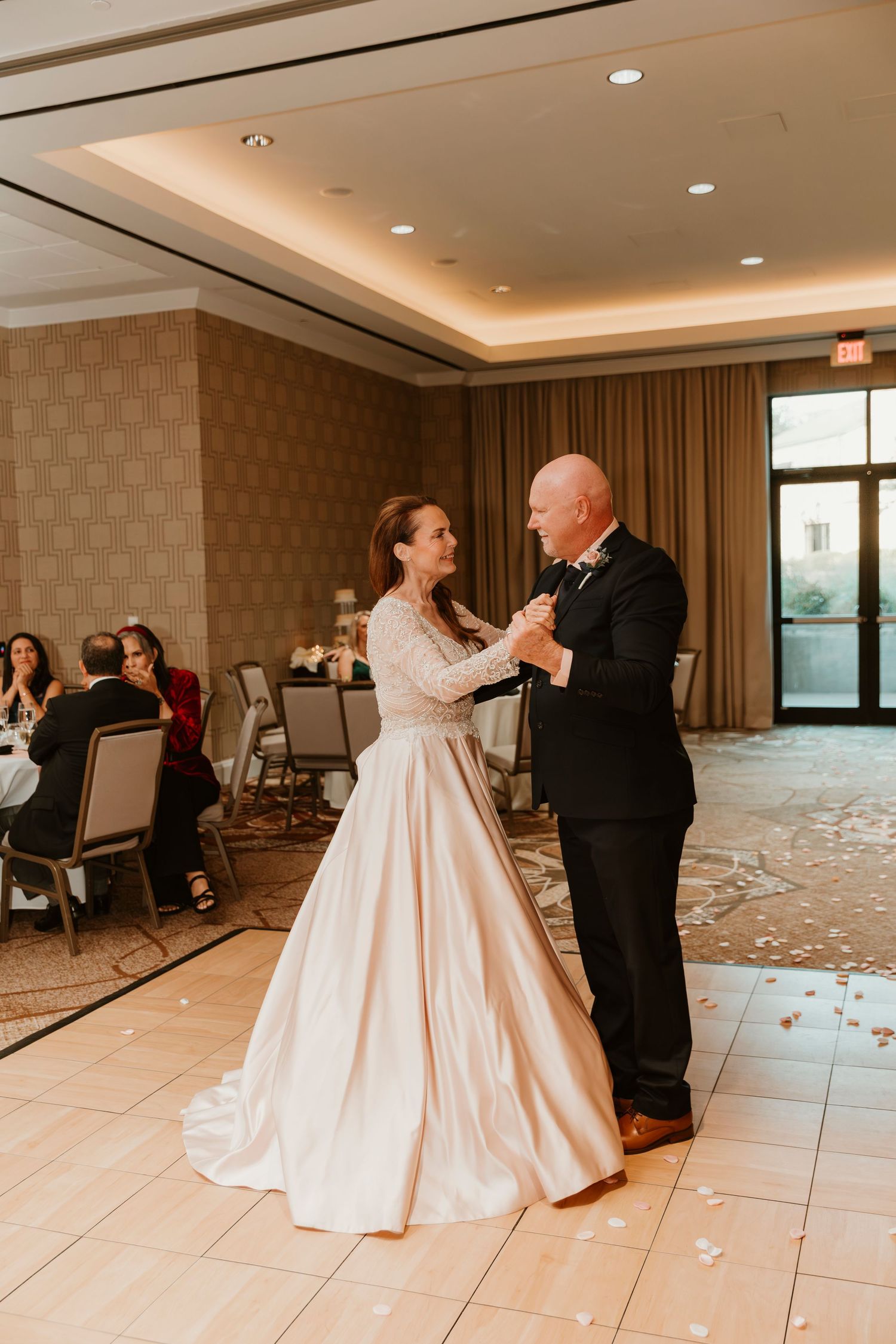 A couple shares their first dance in an elegant ballroom with soft lighting and scattered rose petals on the floor.