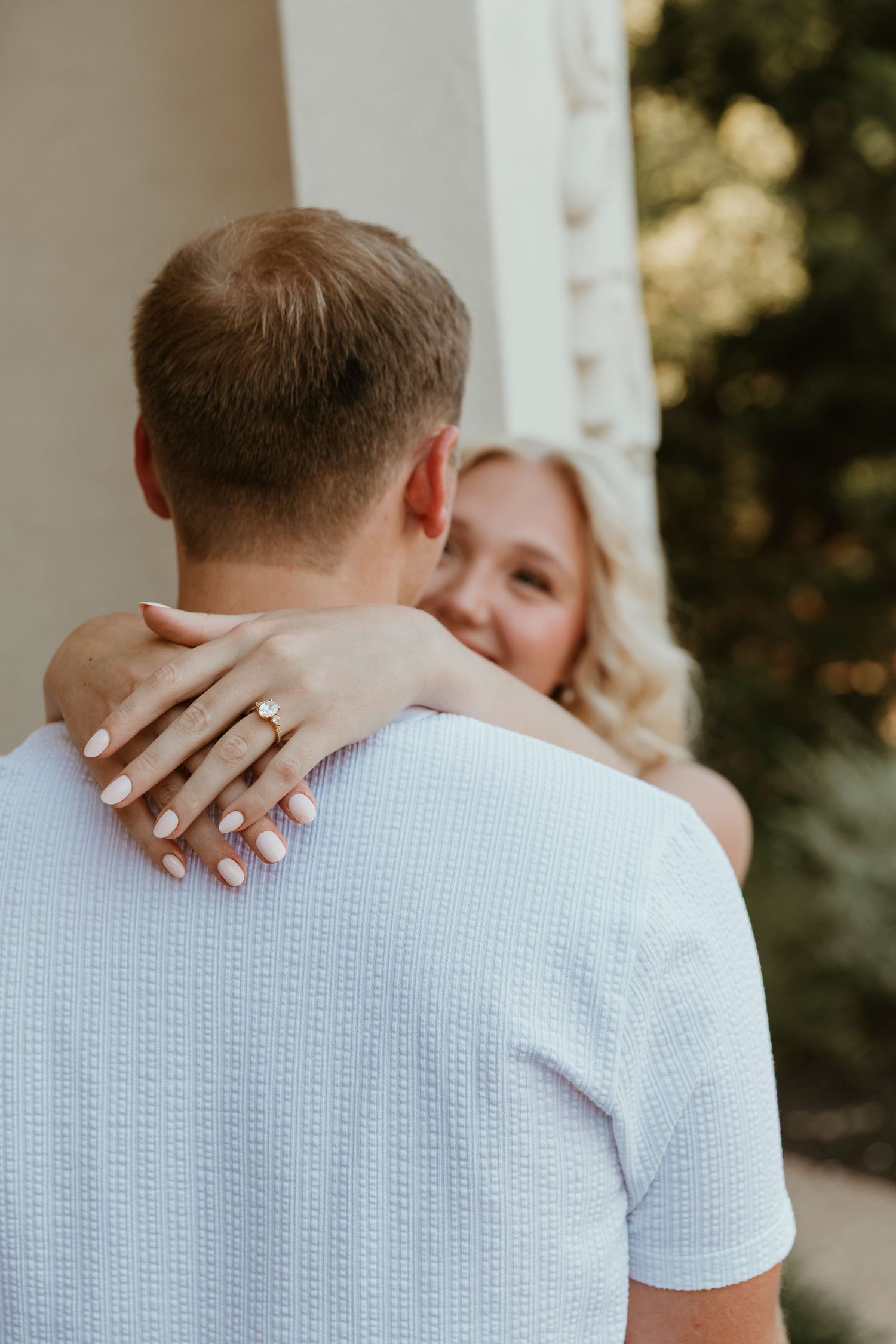 A couple embraces outdoors, with the woman's hands displaying her engagement ring as she hugs her partner.