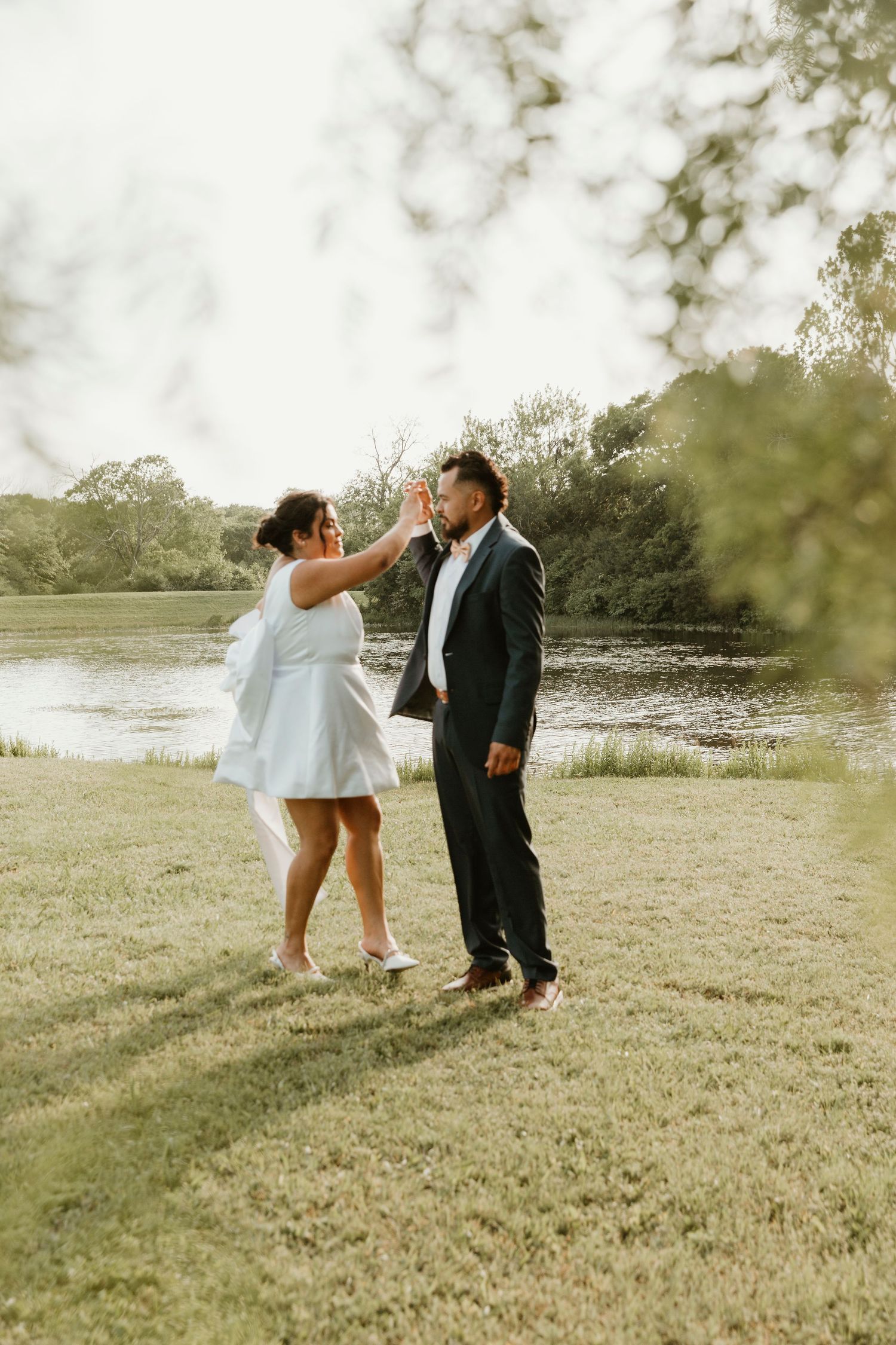 Wedding couple sharing playful moments in a sunlit meadow by a lake.