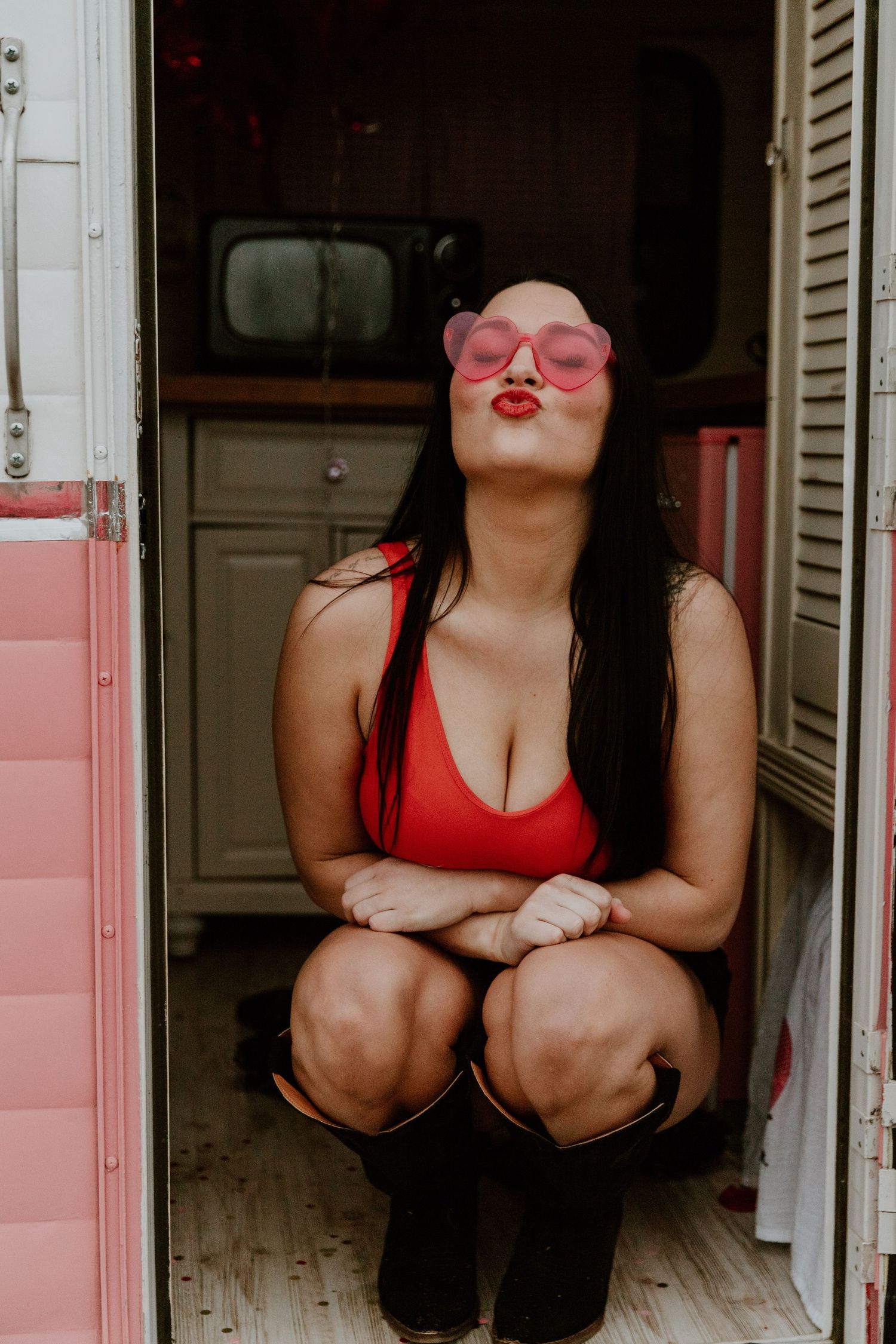 Woman in red tank top and pink sunglasses posing playfully in vintage-style indoor setting.