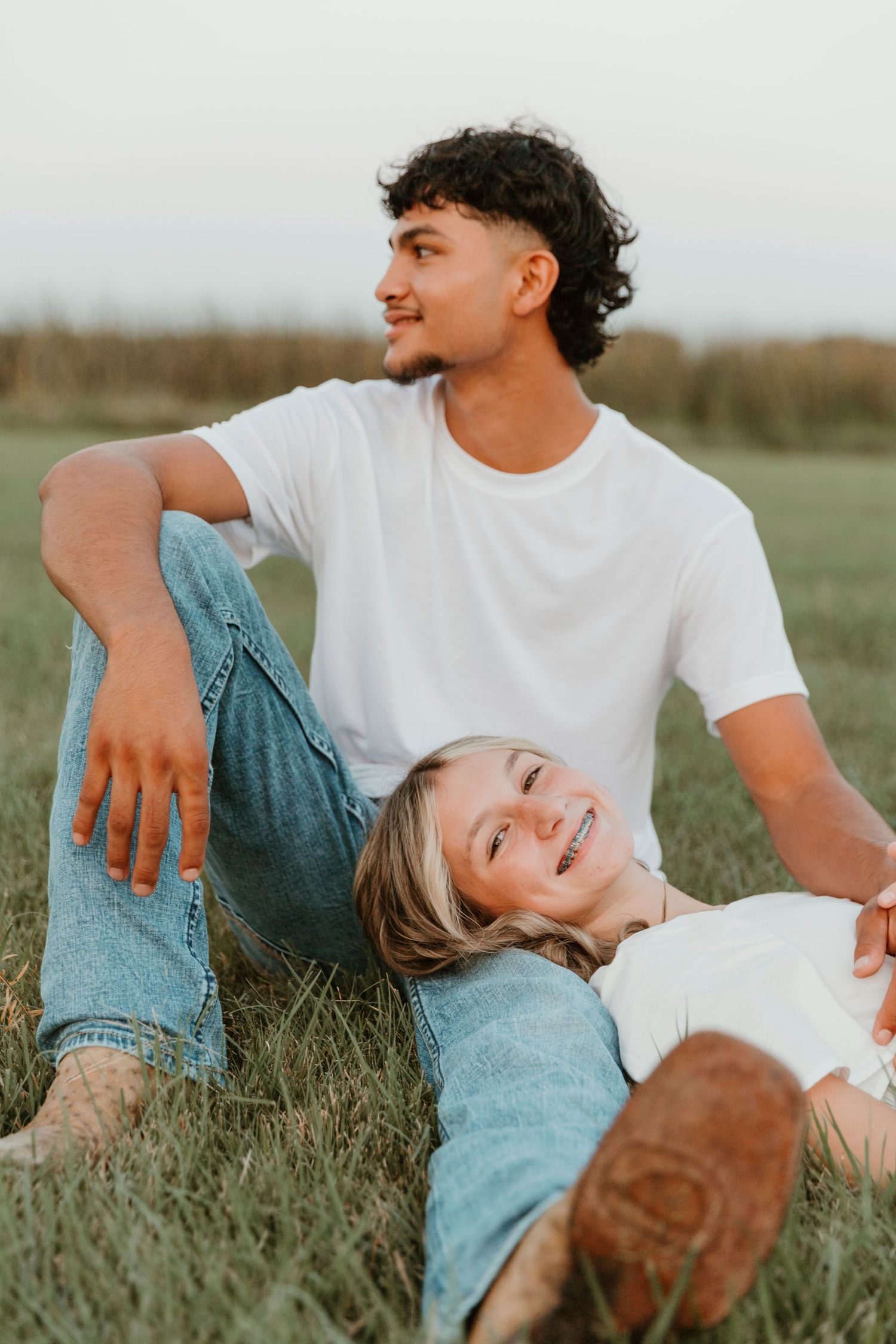 Couple lying in grass field wearing white t-shirts and jeans sharing joyful laughter in warm sunlight.
