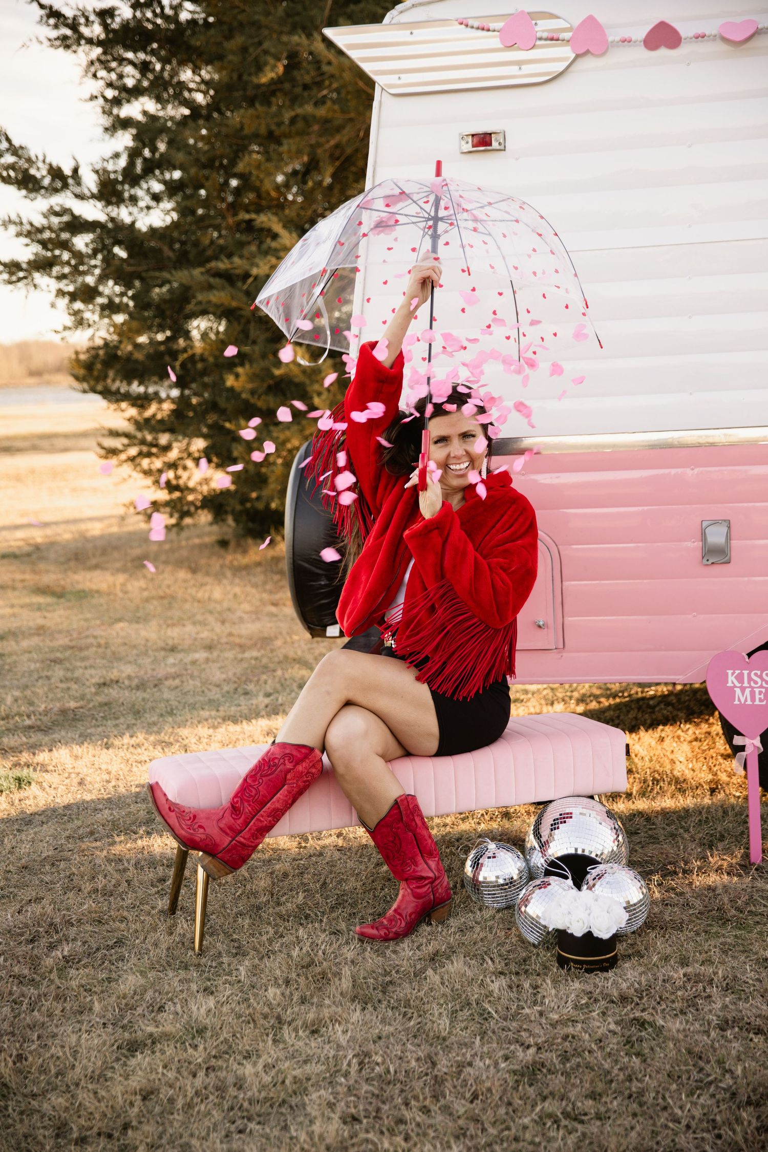 Person in red cardigan and boots sits with pink umbrella next to retro camper.