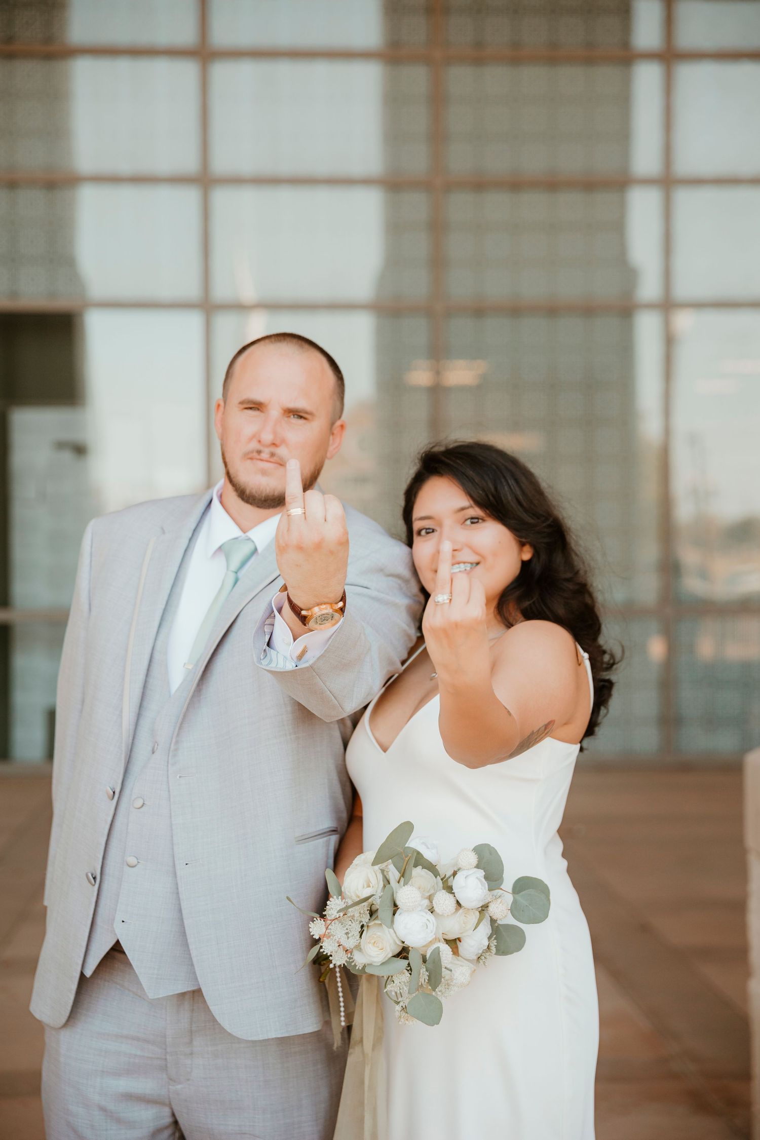 A wedding couple in formal attire making playful gestures at the camera in front of a modern building.