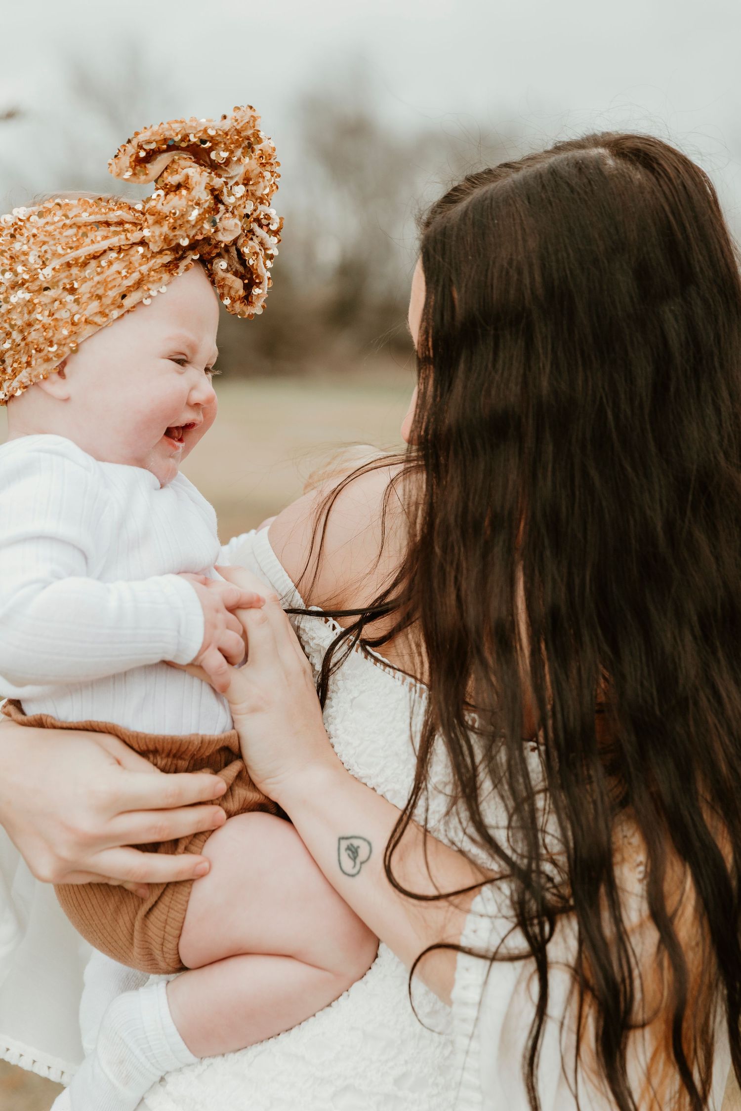 Baby wearing orange bow headband laughing with mother in outdoor setting.