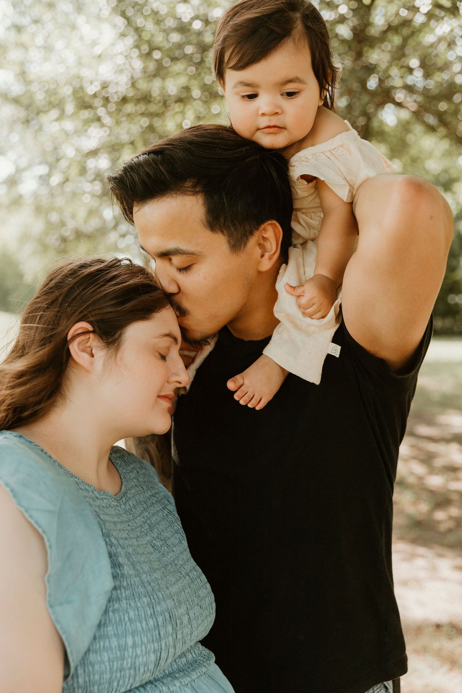 A tender family moment outdoors with parents holding their baby against a sunlit background.