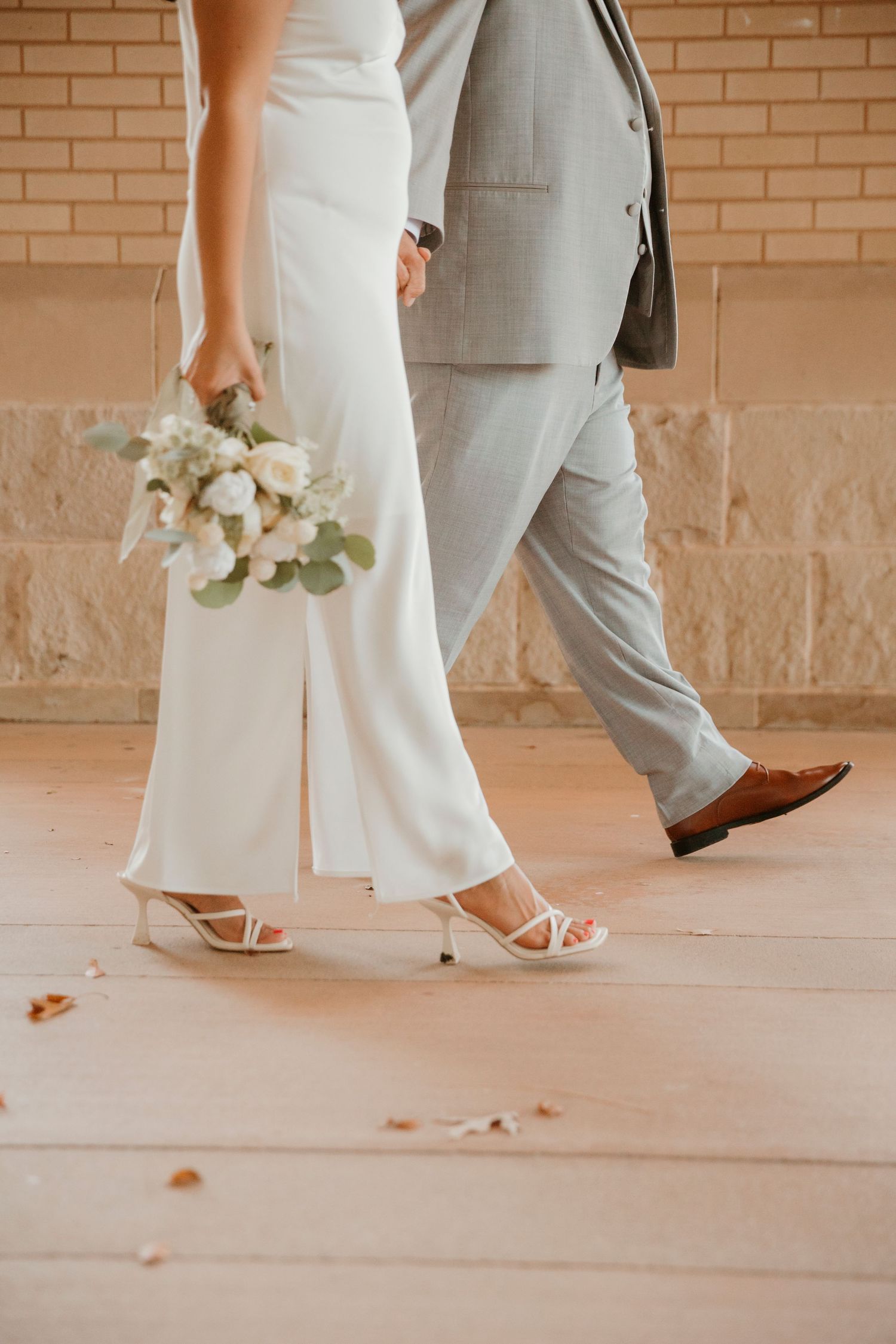 Wedding couple's feet and legs walking together on brick patio with bride holding white floral bouquet.