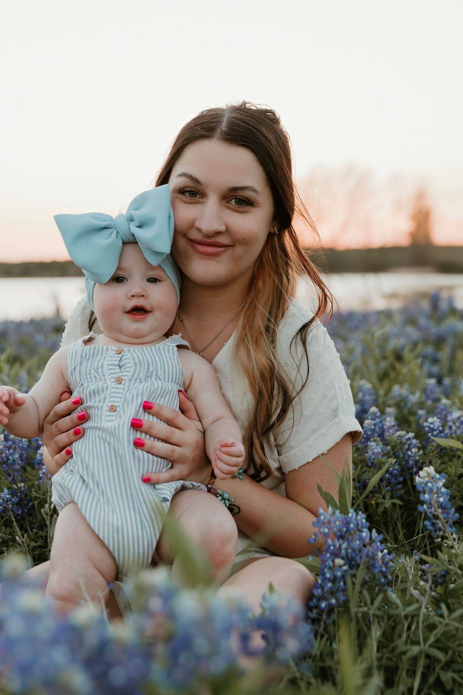 Mother and baby wearing blue bow headband pose together in field of bluebonnets.