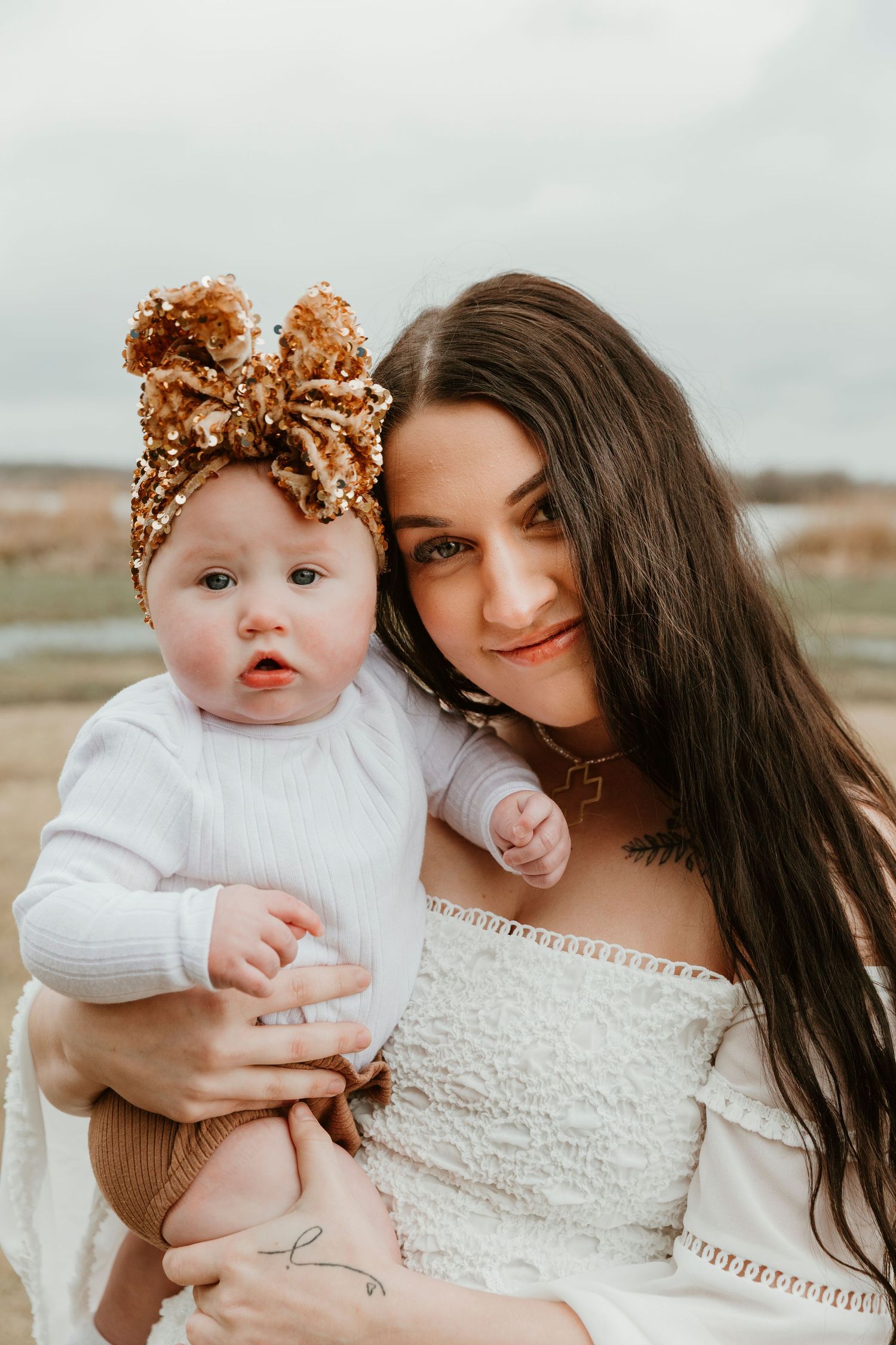 Mother and baby in white outfits pose outdoors, baby wearing gold sequin bow.