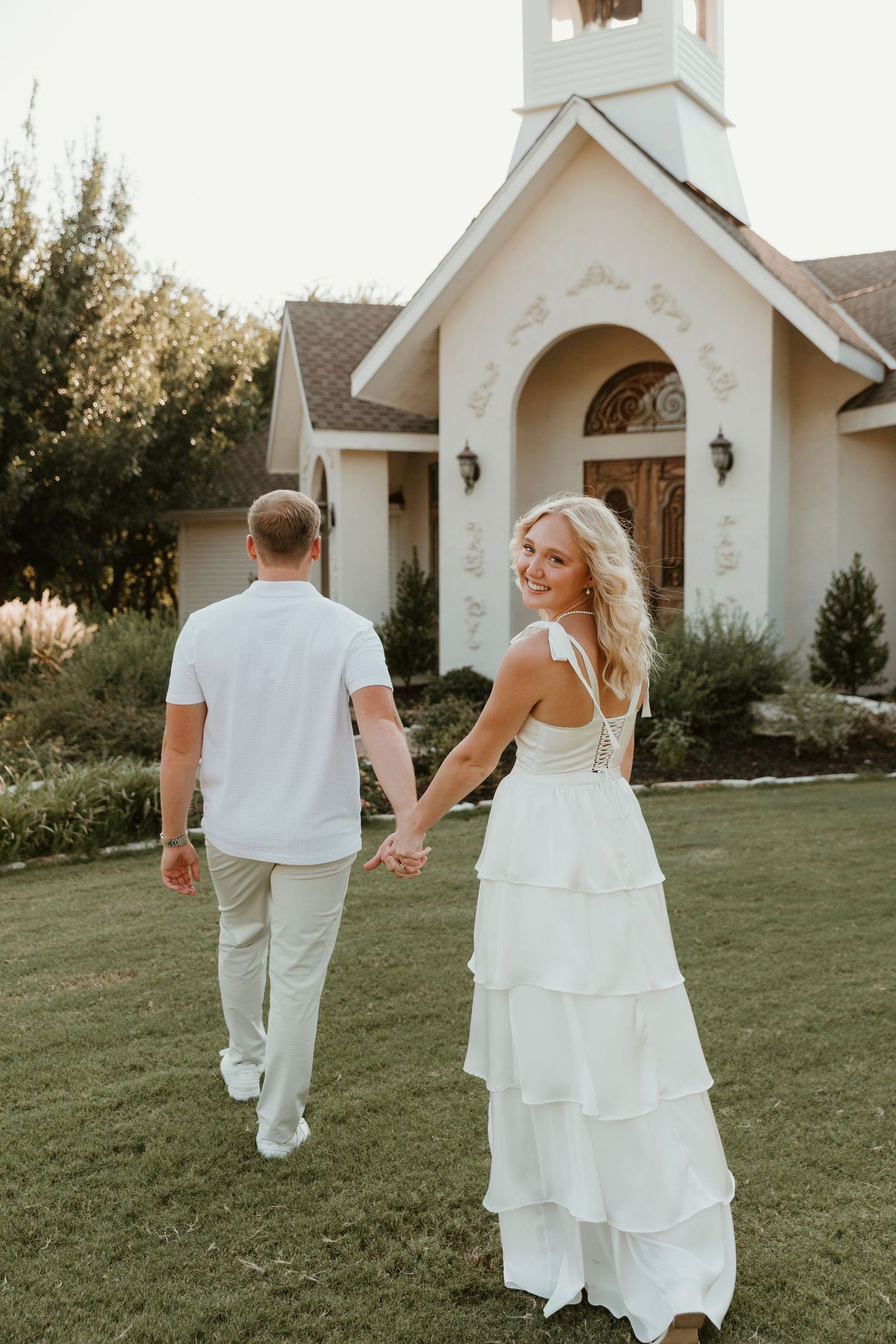 A couple walks hand in hand on a lawn in front of a small white chapel wearing coordinated white and beige outfits.