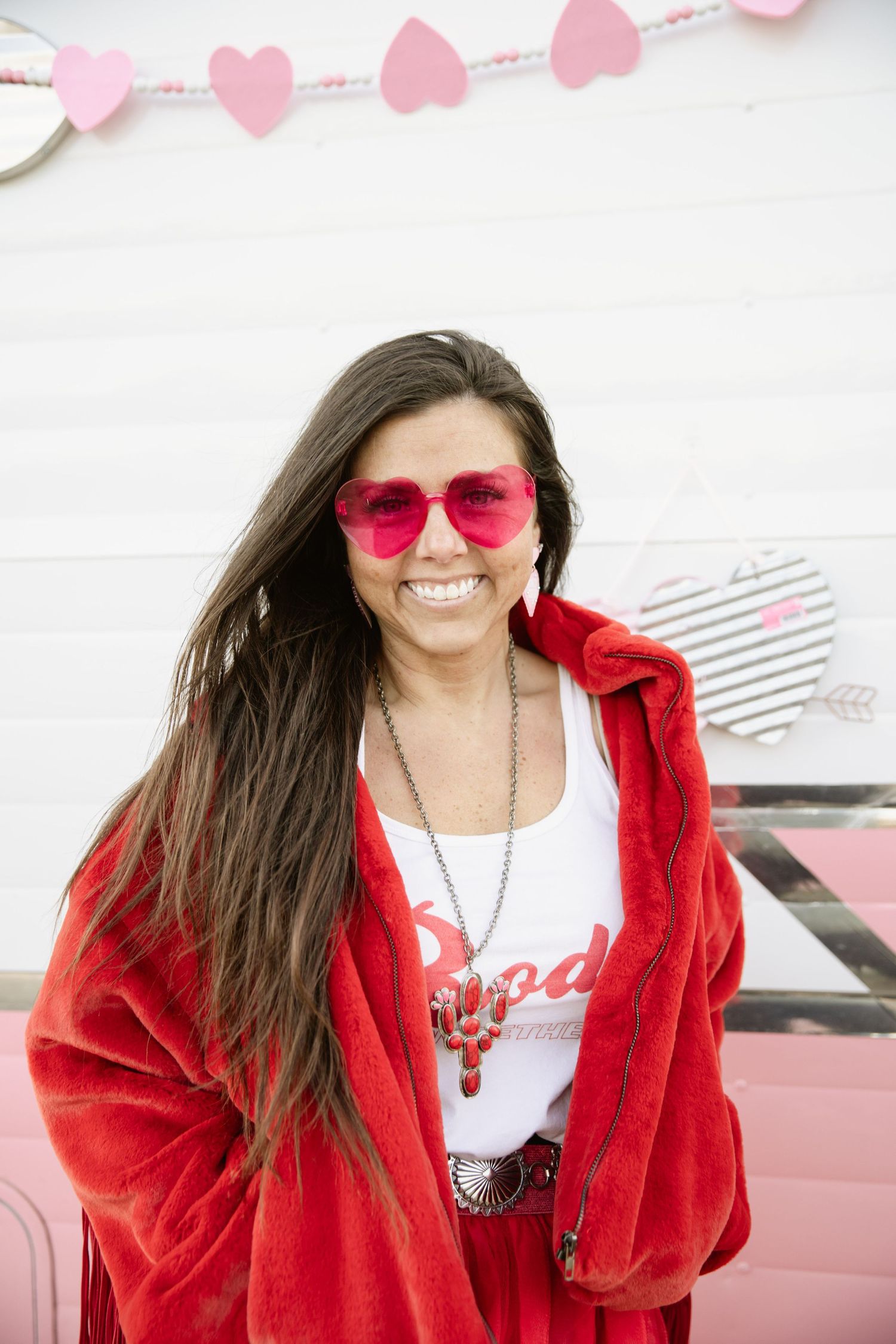 Woman in red jacket and pink sunglasses poses against white wall with heart decorations.