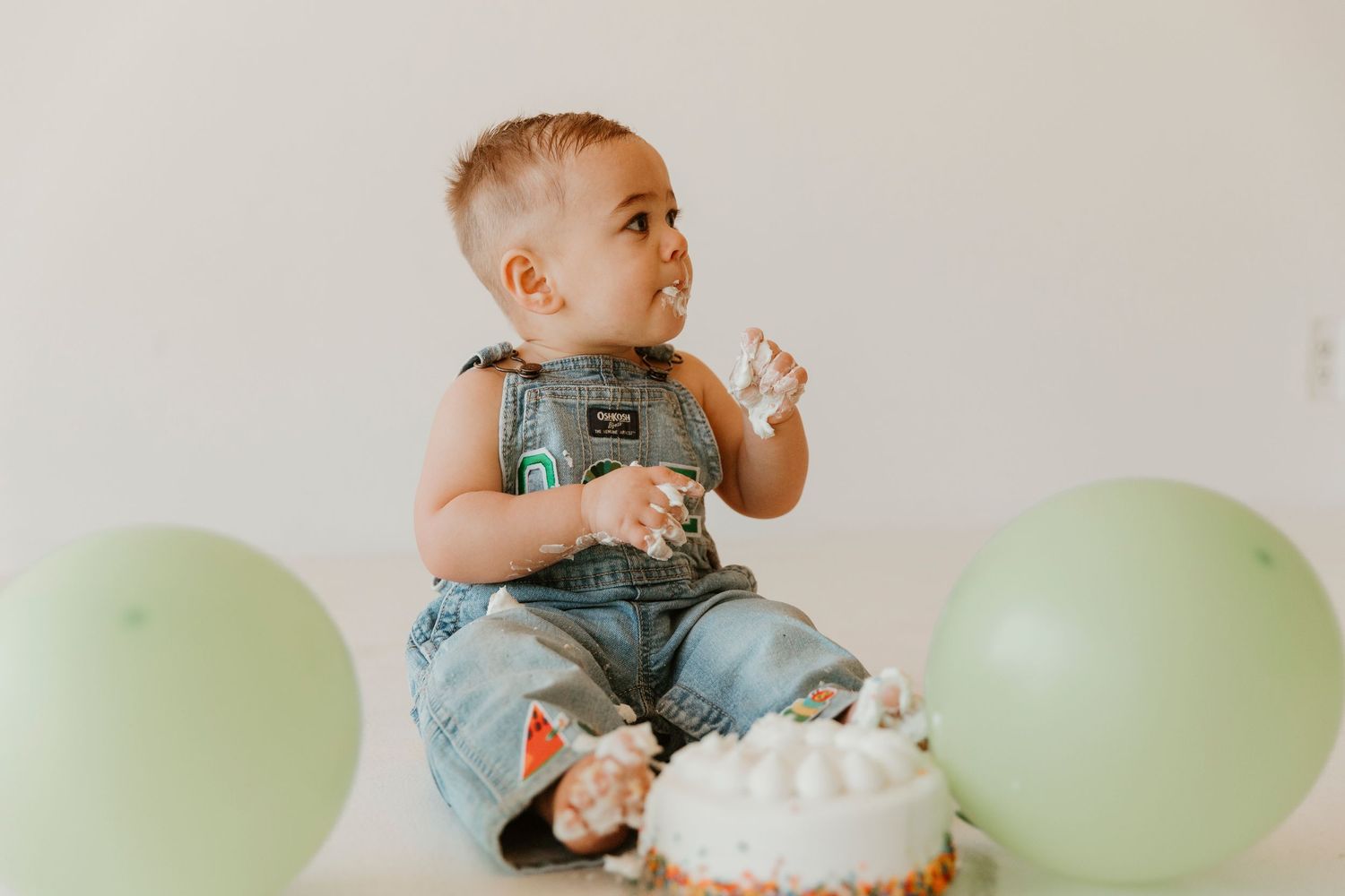 Baby in denim overalls sits on floor with green balloons and white frosted cake for first birthday celebration.