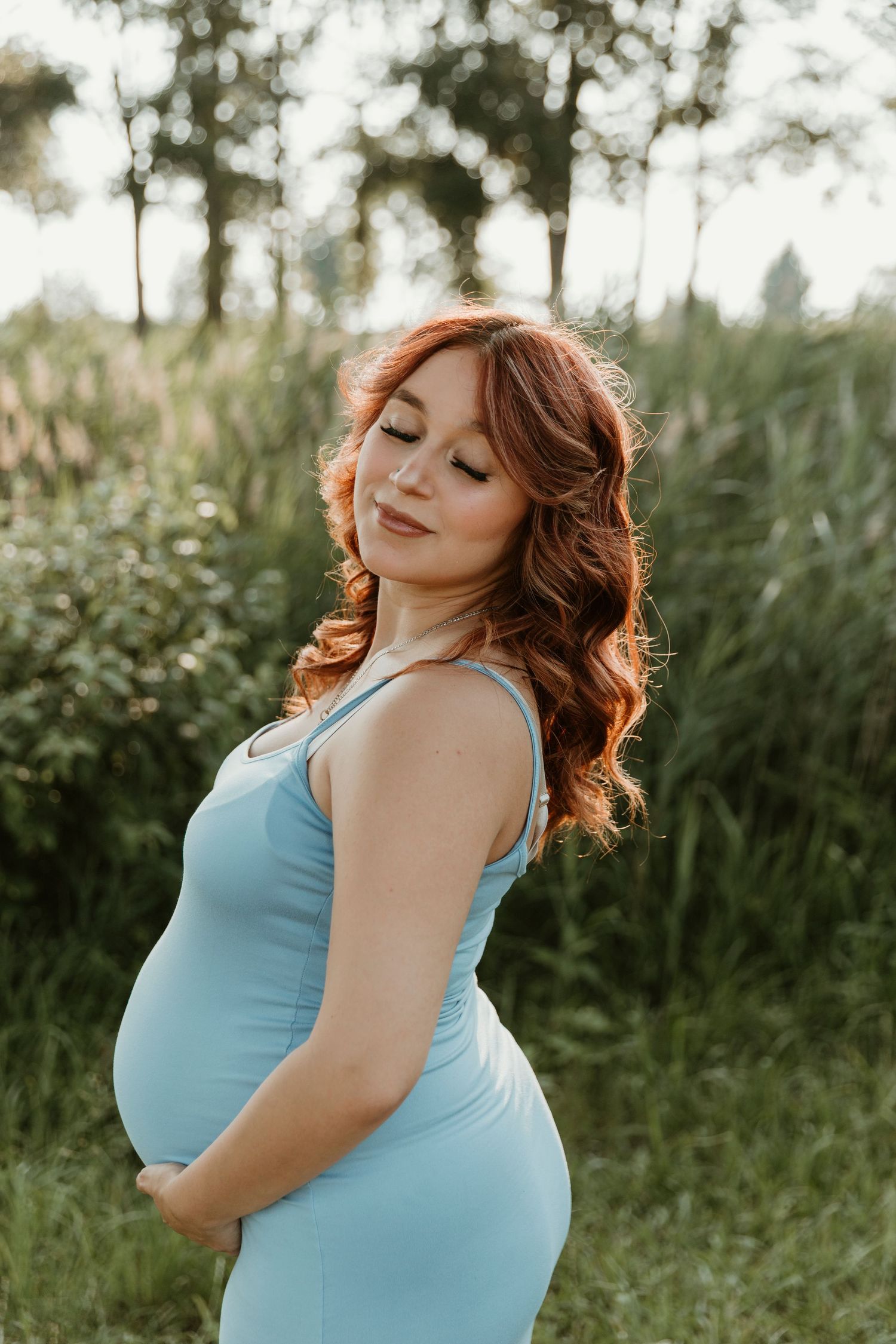 A light blue maternity dress catches golden sunlight in an outdoor photoshoot among tall grass and trees.