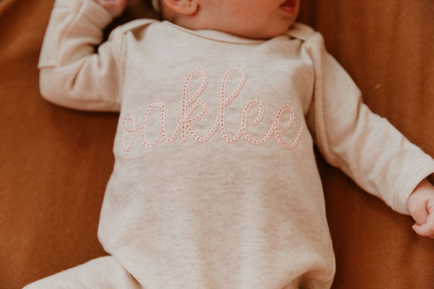 Close up of baby wearing cream colored sweater with text embroidery lying on brown fabric.