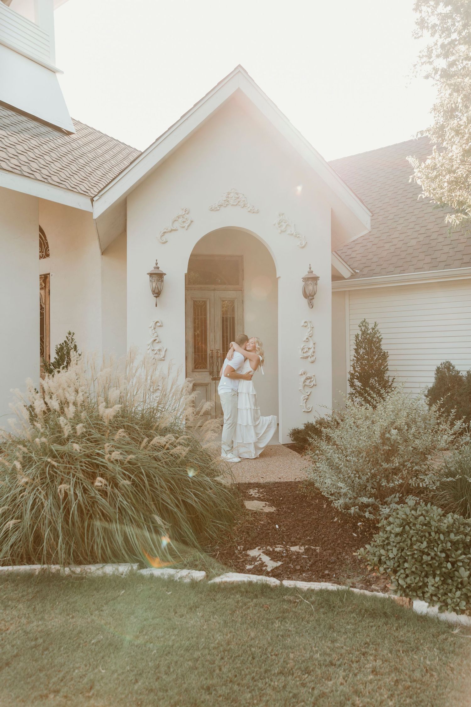 A romantic couple embraces under an arched church entrance surrounded by ornate landscaping at sunset.