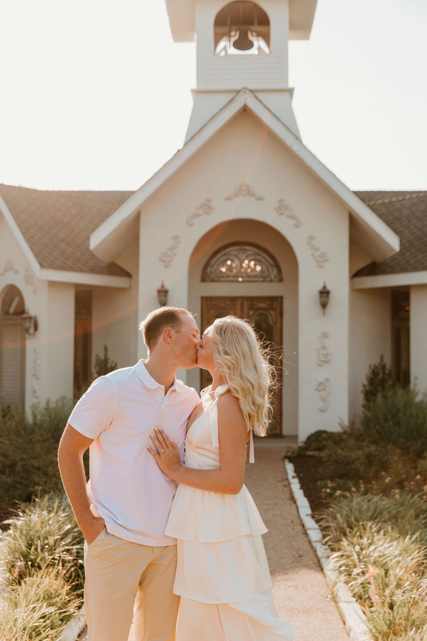A couple shares a romantic moment in front of a charming white chapel at sunset during a golden hour photoshoot.