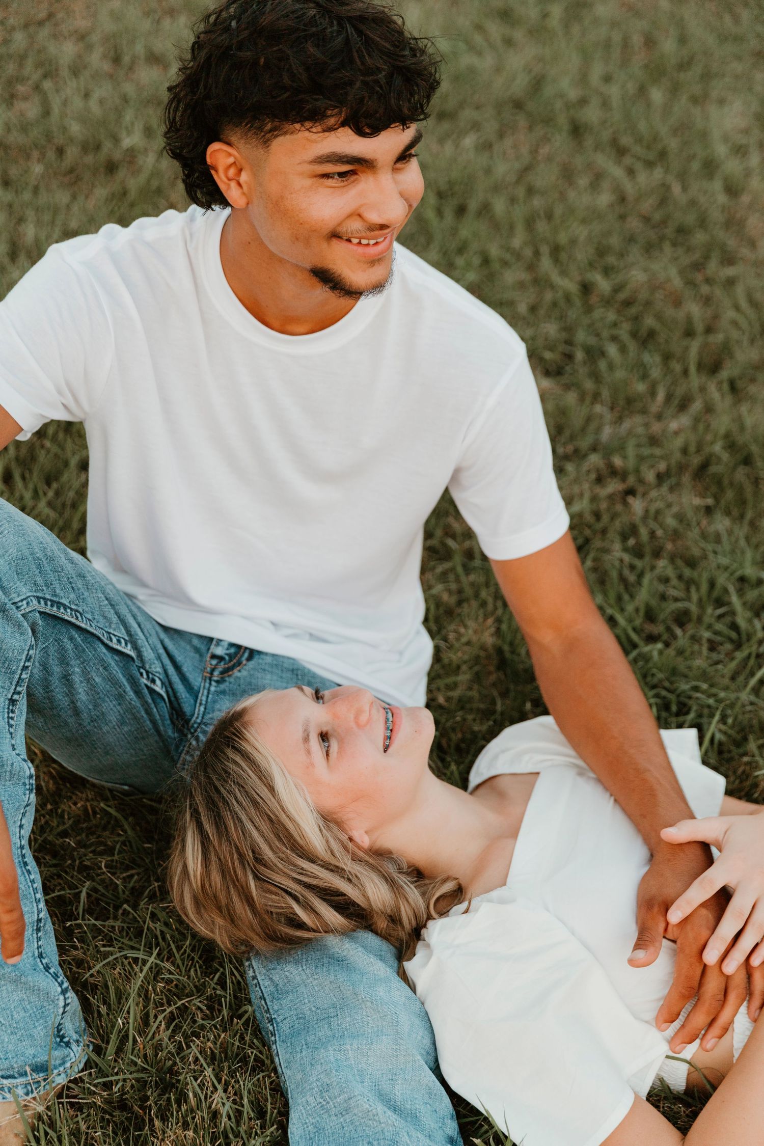 Couple in casual attire sharing a playful moment while lying on grass in warm sunlight.