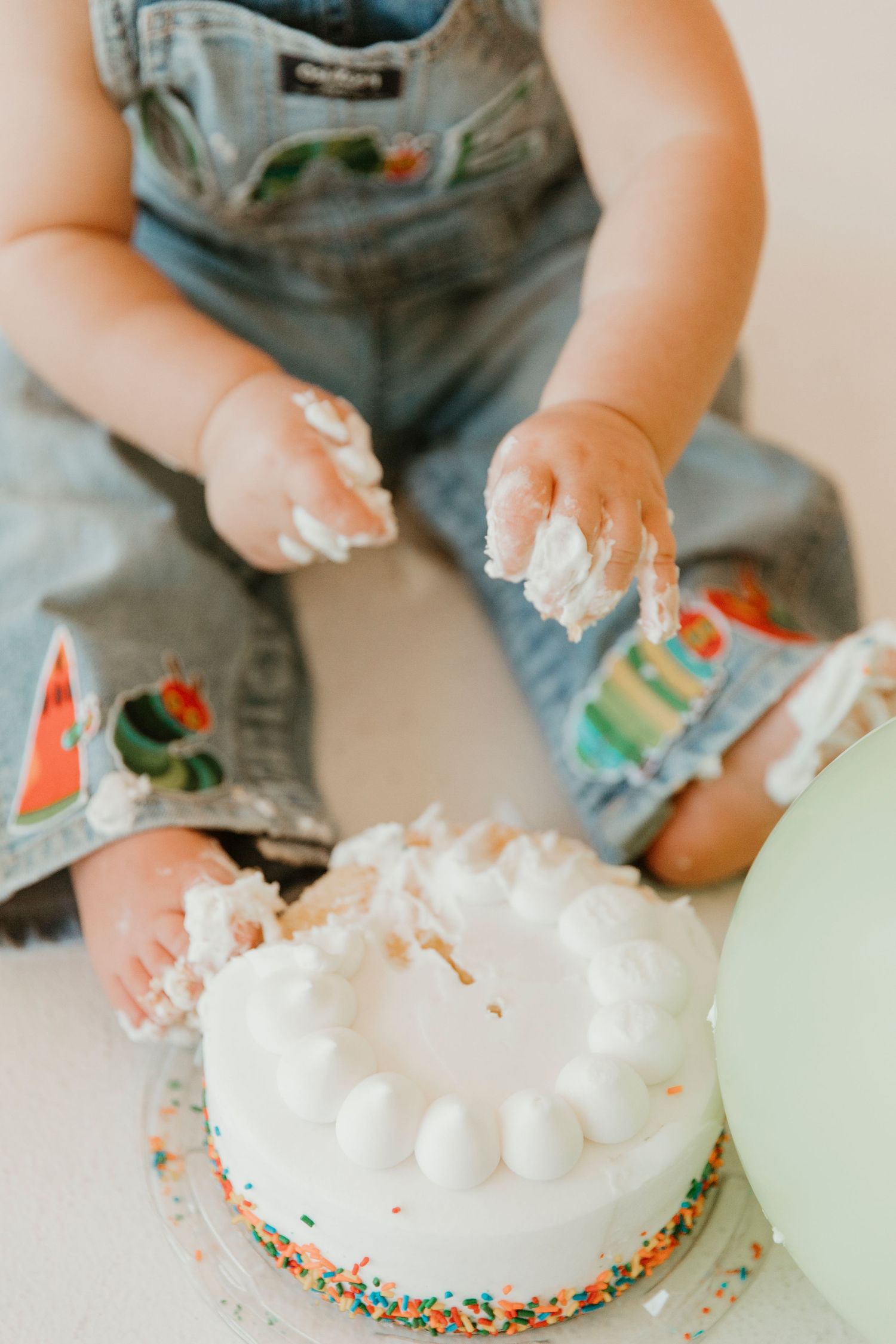 Small hands covered in white frosting reach for a decorated birthday cake with sprinkles.