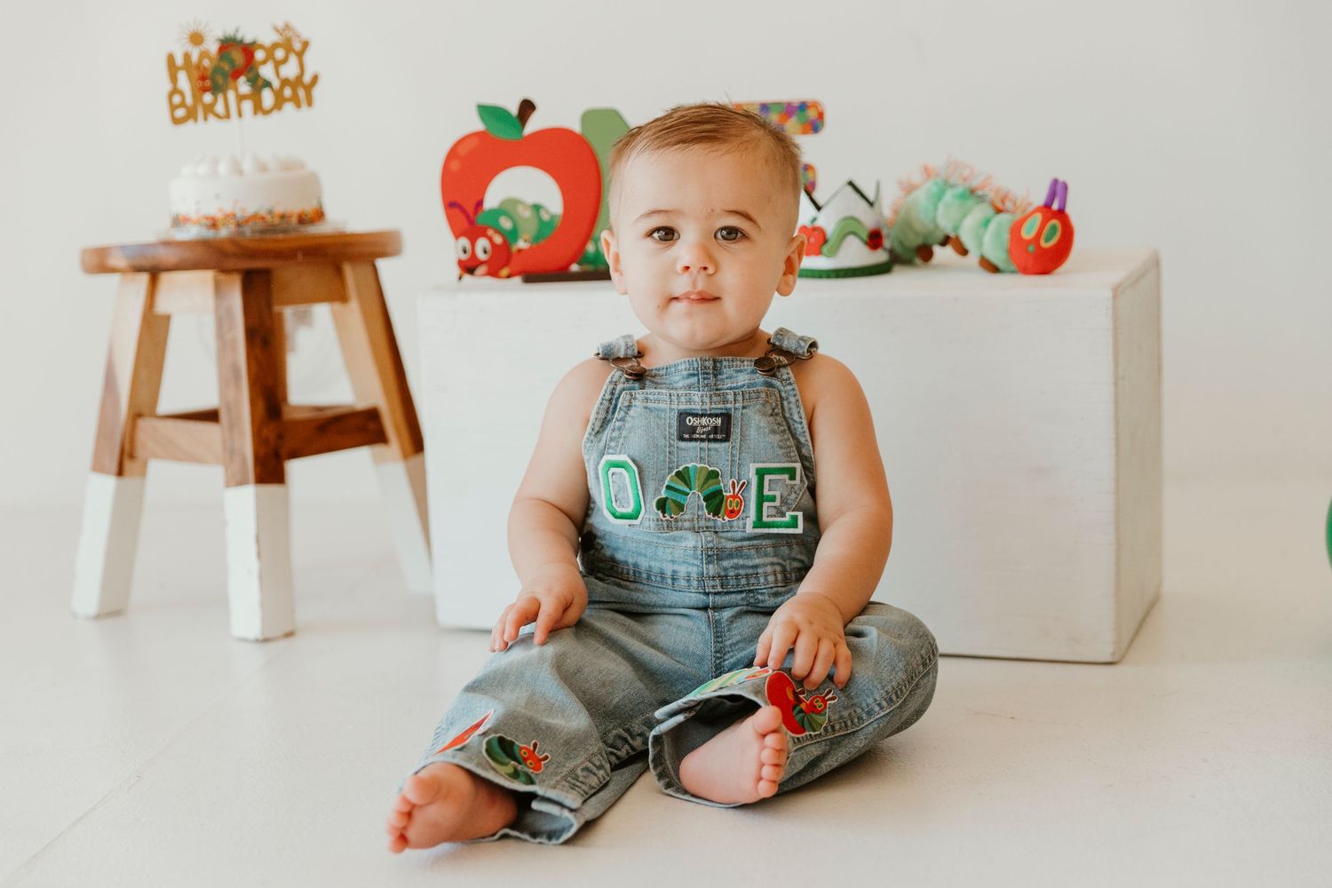 A small child wearing green overalls sits on a white floor with holiday decorations visible in the background.
