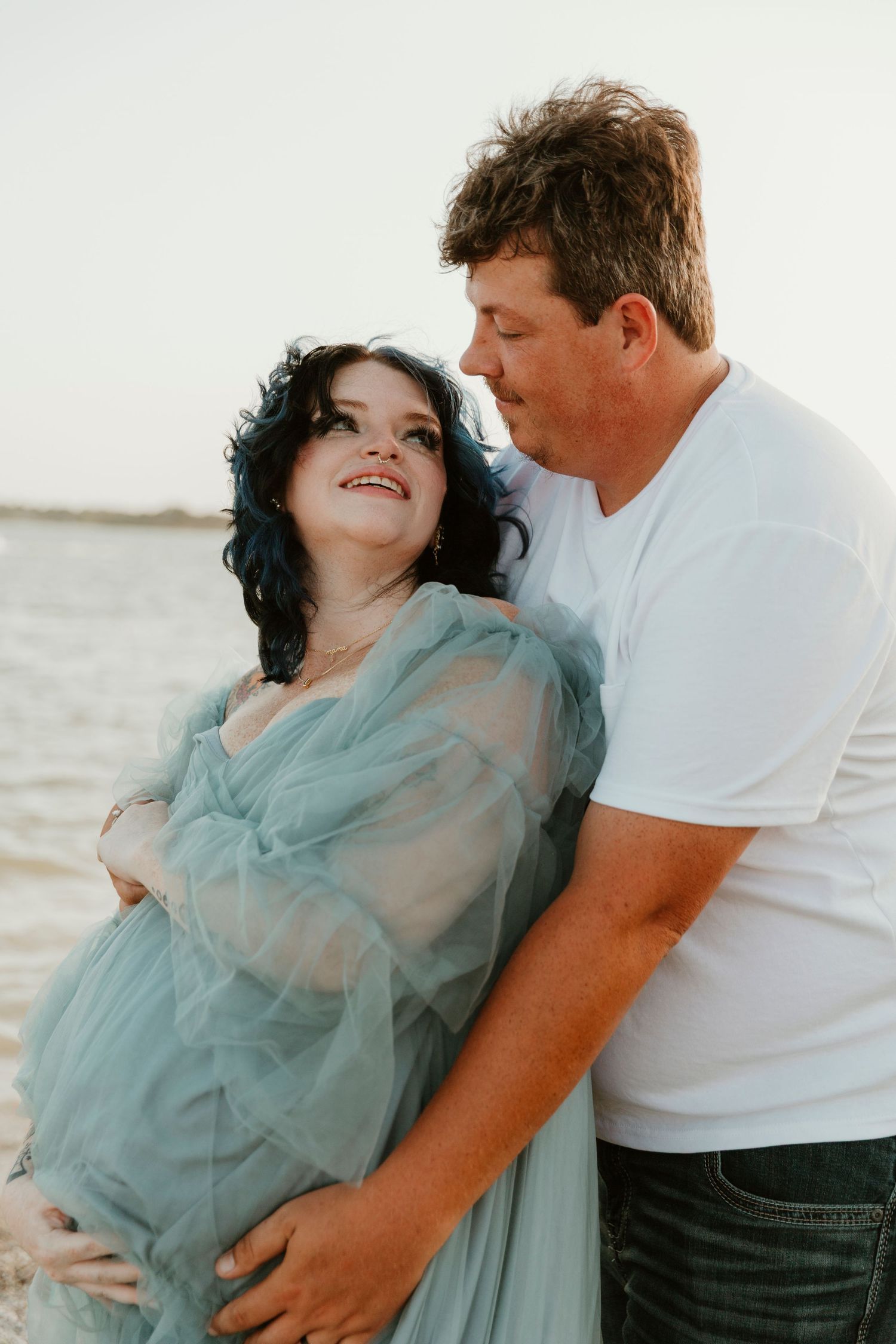 A couple embraces on the beach at sunset, wearing coordinating blue and white outfits.