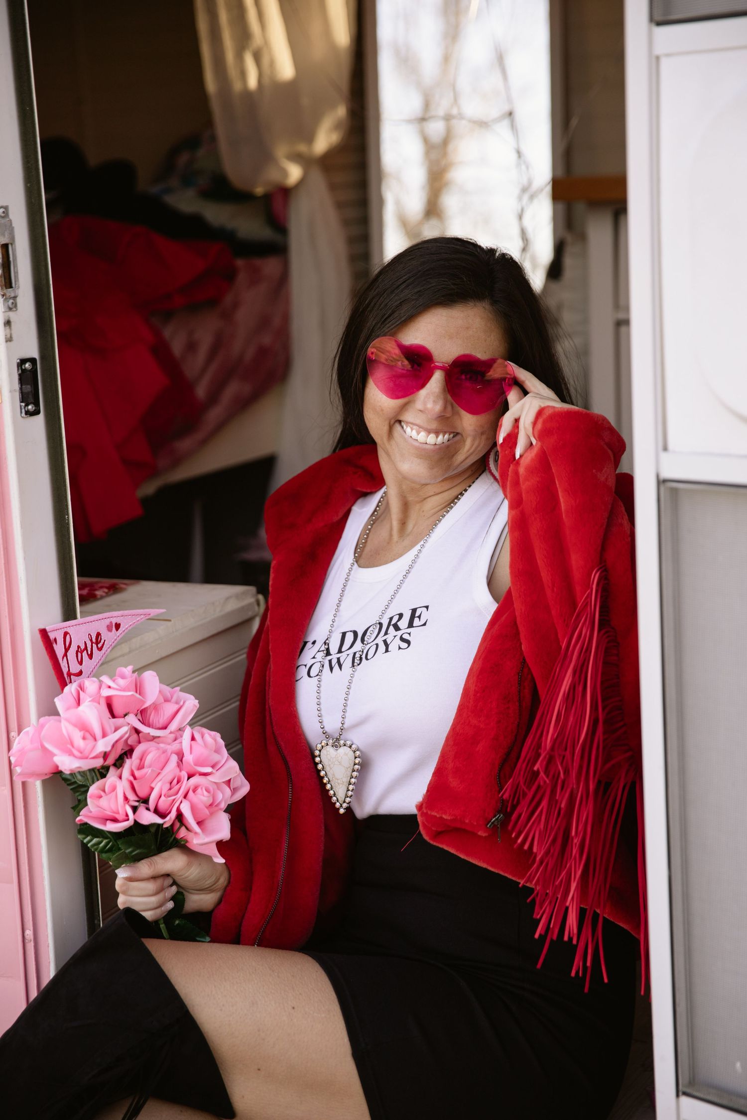 Woman in red fringe cardigan and heart sunglasses holds pink flowers in doorway.