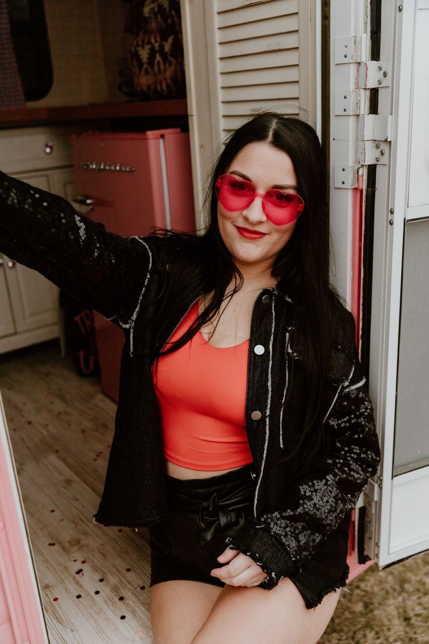 Fashion portrait of woman in red tank top and black sequined jacket posing near vintage pink fridge.