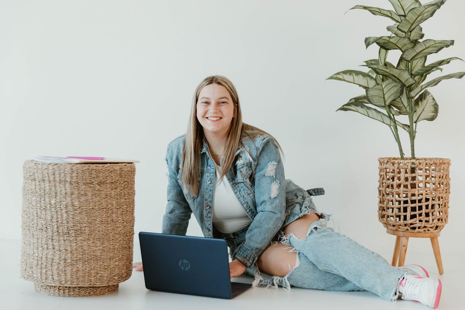 Person in denim jacket working on laptop with decorative plant and basket in minimalist setting.