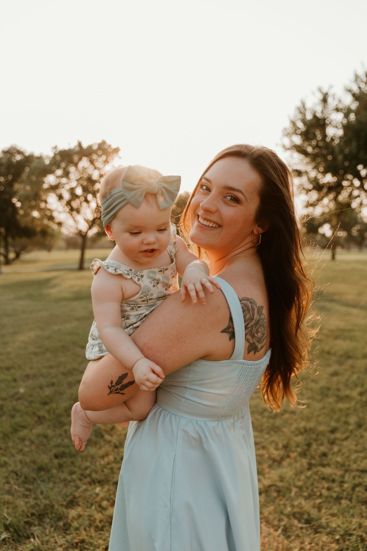 A woman in a light blue dress holds a baby at sunset in a grassy field.
