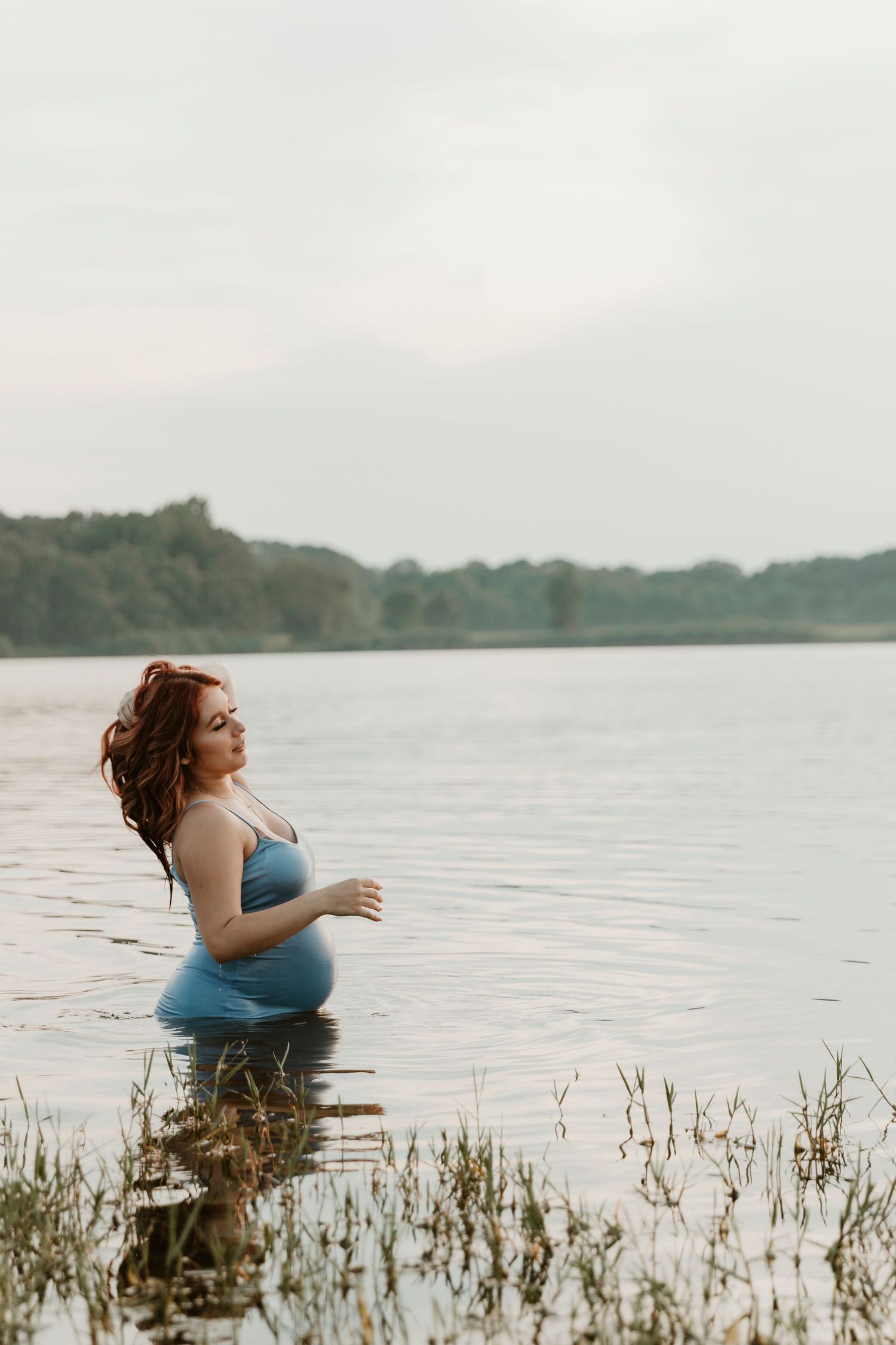 Expectant mother in a blue dress poses gracefully in a serene lake setting during golden hour.