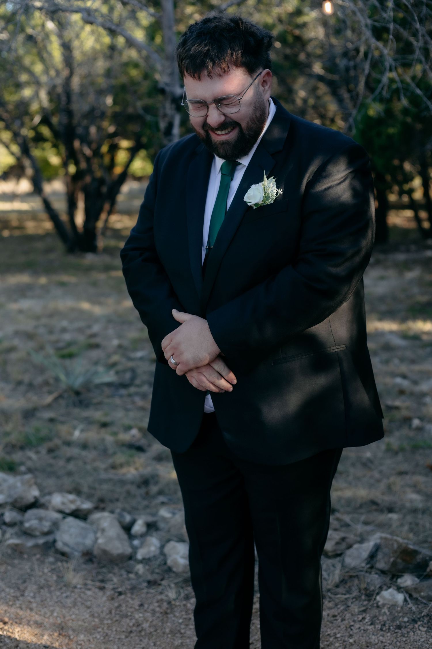 Groom in black suit smiles joyfully during outdoor wedding.