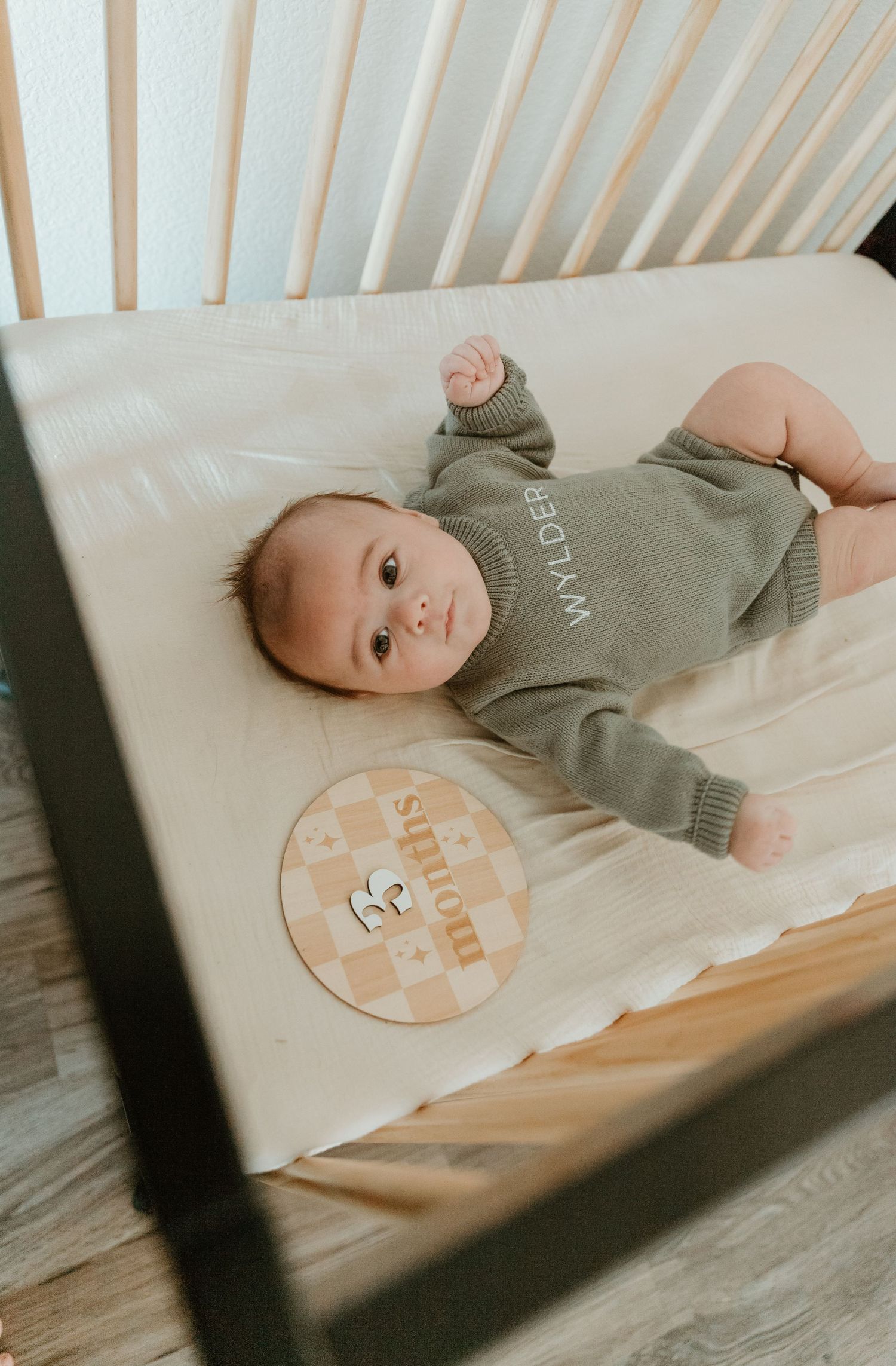 Baby wearing gray onesie lying in white crib with milestone achievement sticker.