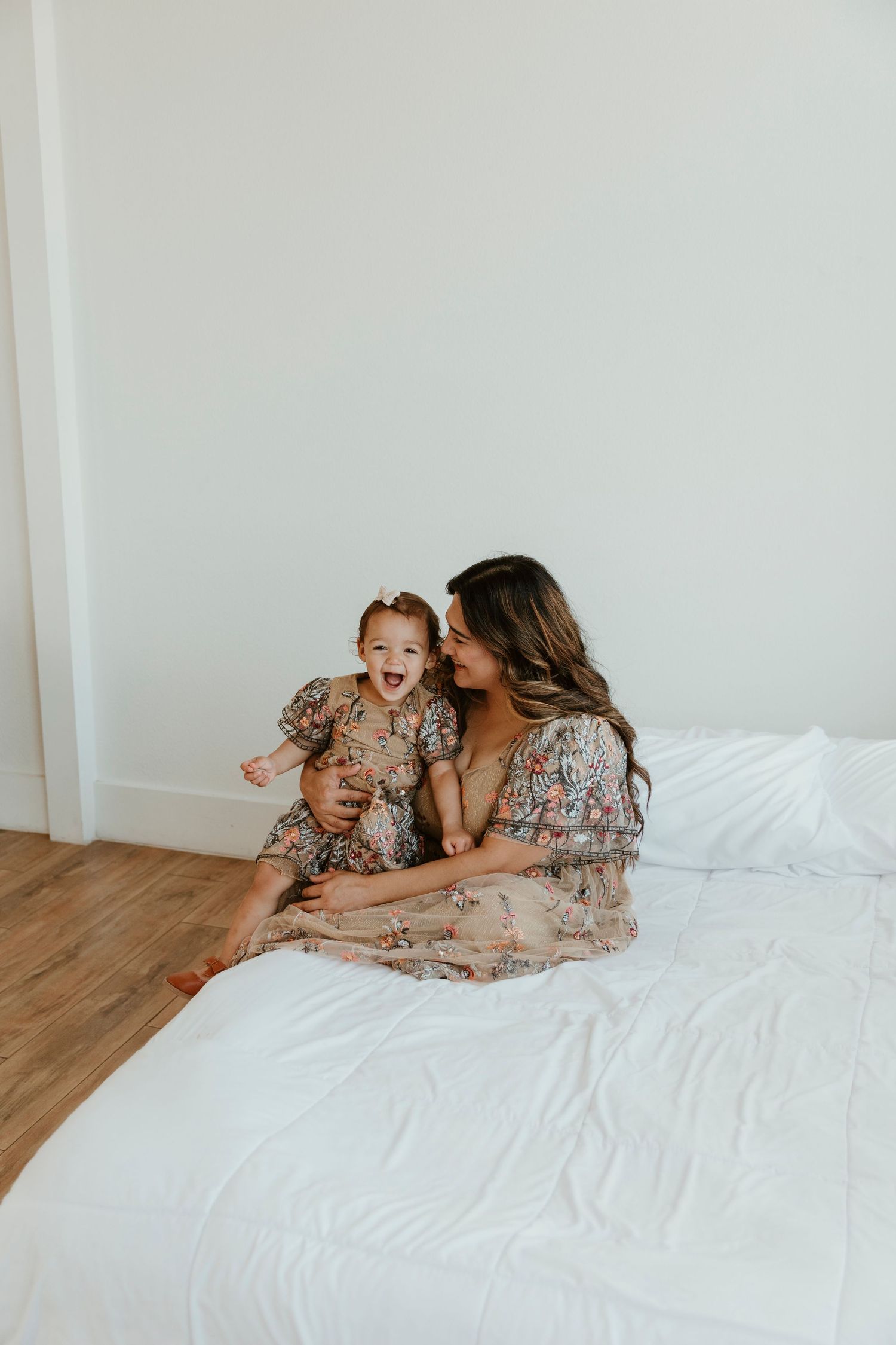 Mother and daughter laughing together on white bedding in matching floral outfits.