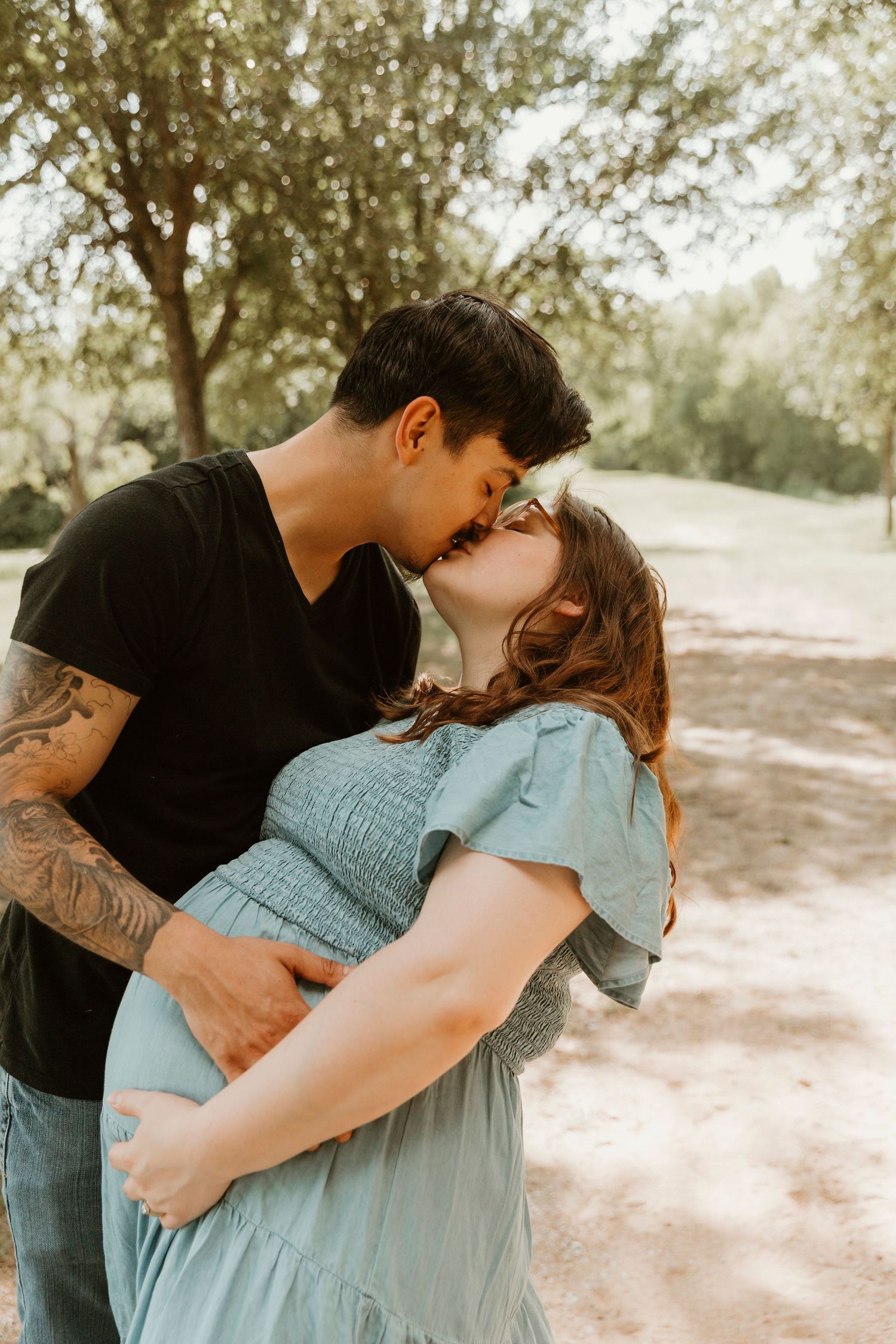 Expecting couple embraces tenderly outdoors, with the pregnant partner wearing a flowing blue dress.