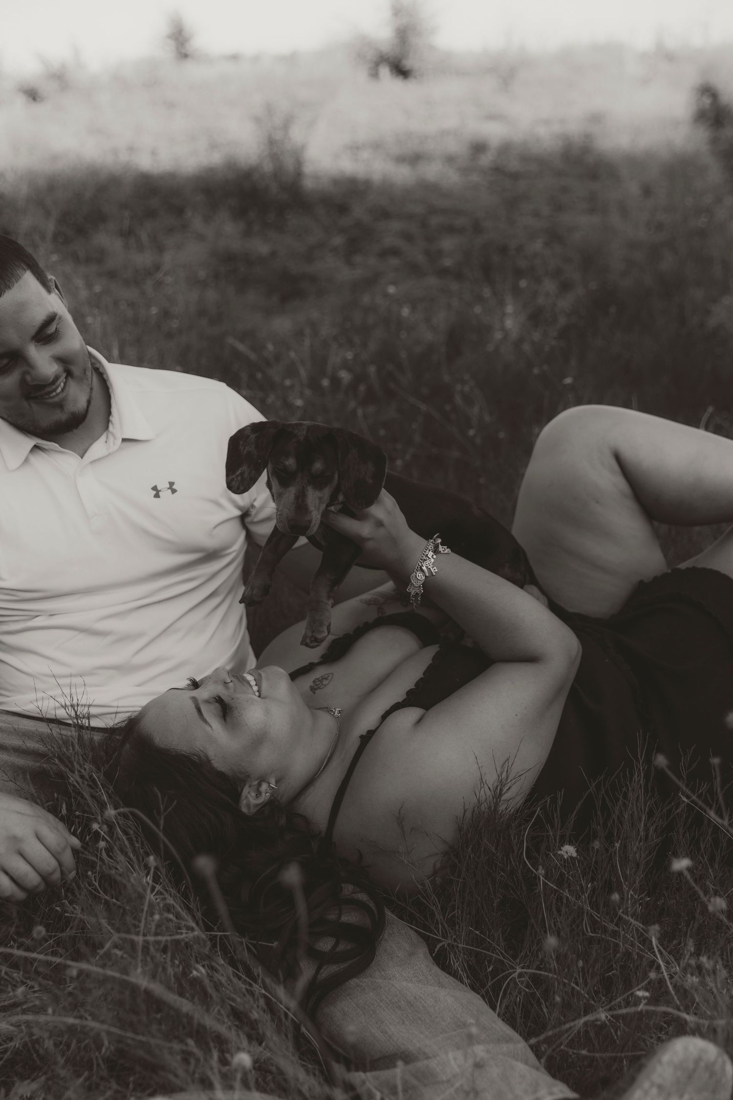 A couple lies in a grassy field in a relaxed black and white portrait.